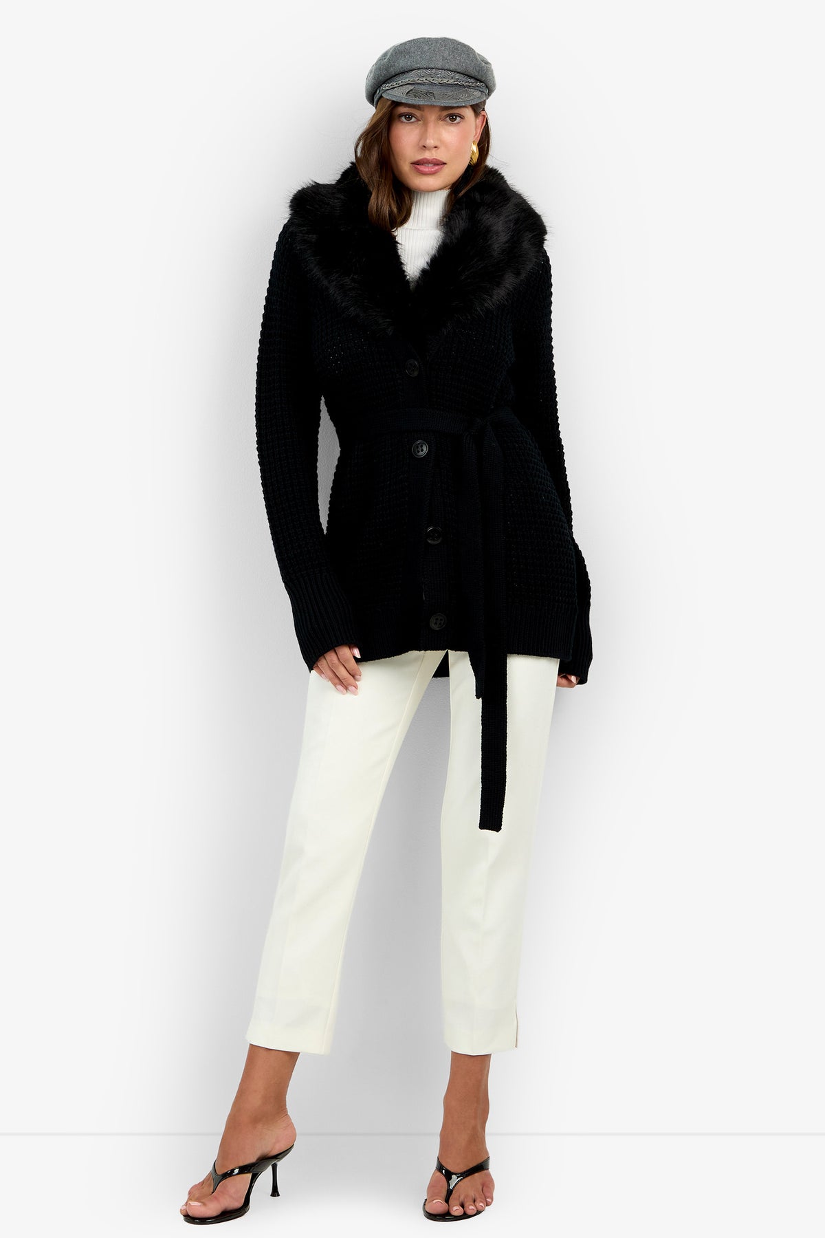 Woman wearing a gray newsboy cap, the Pellicia Fur Cardigan in black with a belted waist and faux fur collar, a white turtleneck, white cropped pants, and black sandals standing against a white background.