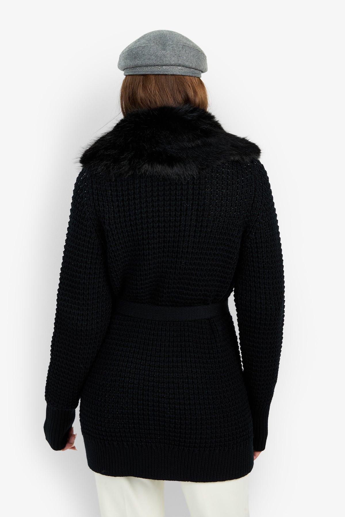 A person seen from the back against a white background is wearing a grey beret, cream pants, and the Pellicia Fur Cardigan—a black textured knit sweater featuring a black faux fur collar.