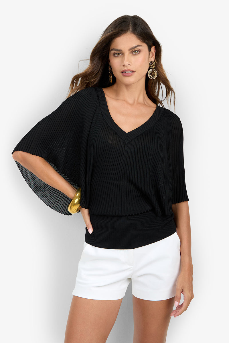 A woman wearing the Nanette Pullover—a black V-neck, short-sleeved, pleated top—paired with white shorts, gold earrings, and a gold bracelet, poses against a plain white background.