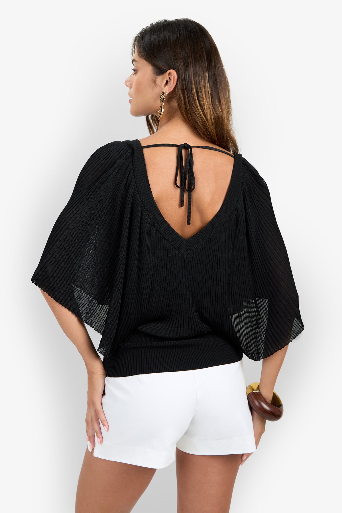 A woman stands facing away with her hand on her hip, showcasing the open-back detail of the black Nanette Pullover—a V-back, tie-neck, pleated top with short sleeves—paired with white shorts.
