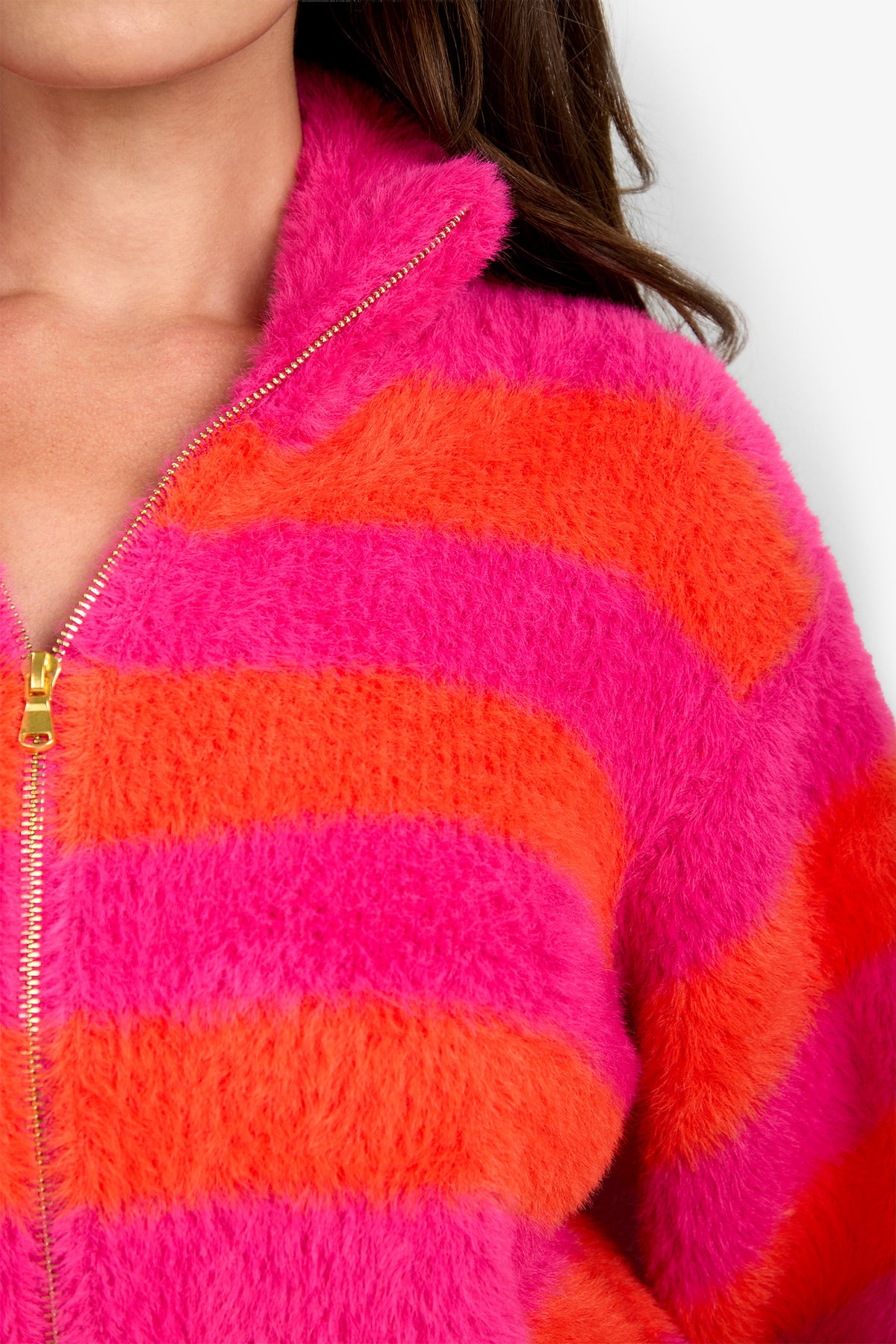 A person wearing the Raja Sweater, which features bold pink and orange horizontal stripes, a fuzzy texture, and a gold zipper, shown from the chest up.