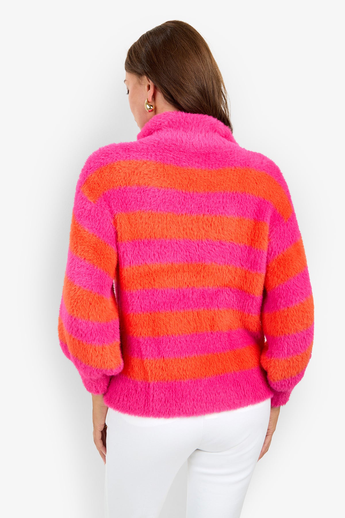 A person with long brown hair faces away from the camera against a white background, wearing the Raja Sweater—a fuzzy knit featuring bold pink and orange horizontal stripes—paired with white pants.
