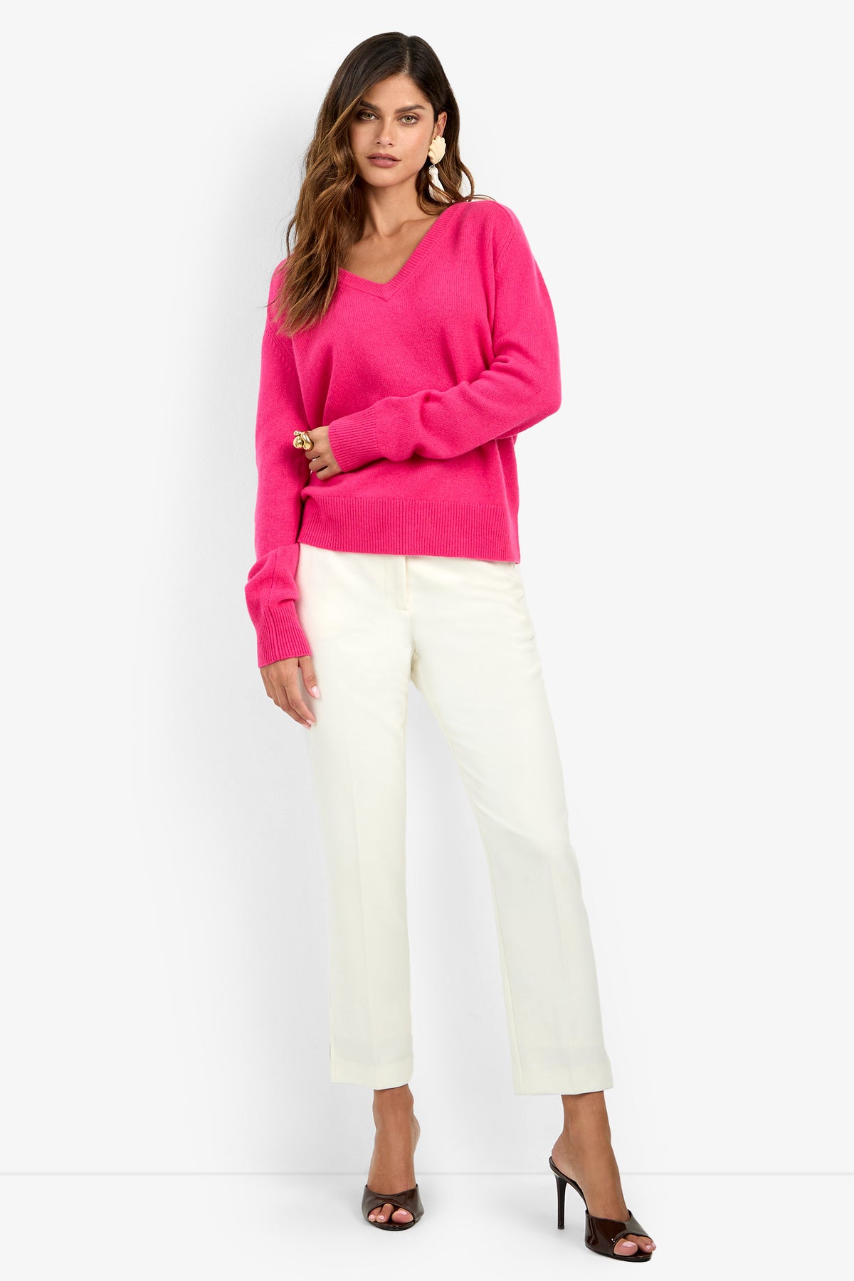 A woman wears a bright pink V-neck EVANGELINE SWEATER with white trousers and black heeled mules.