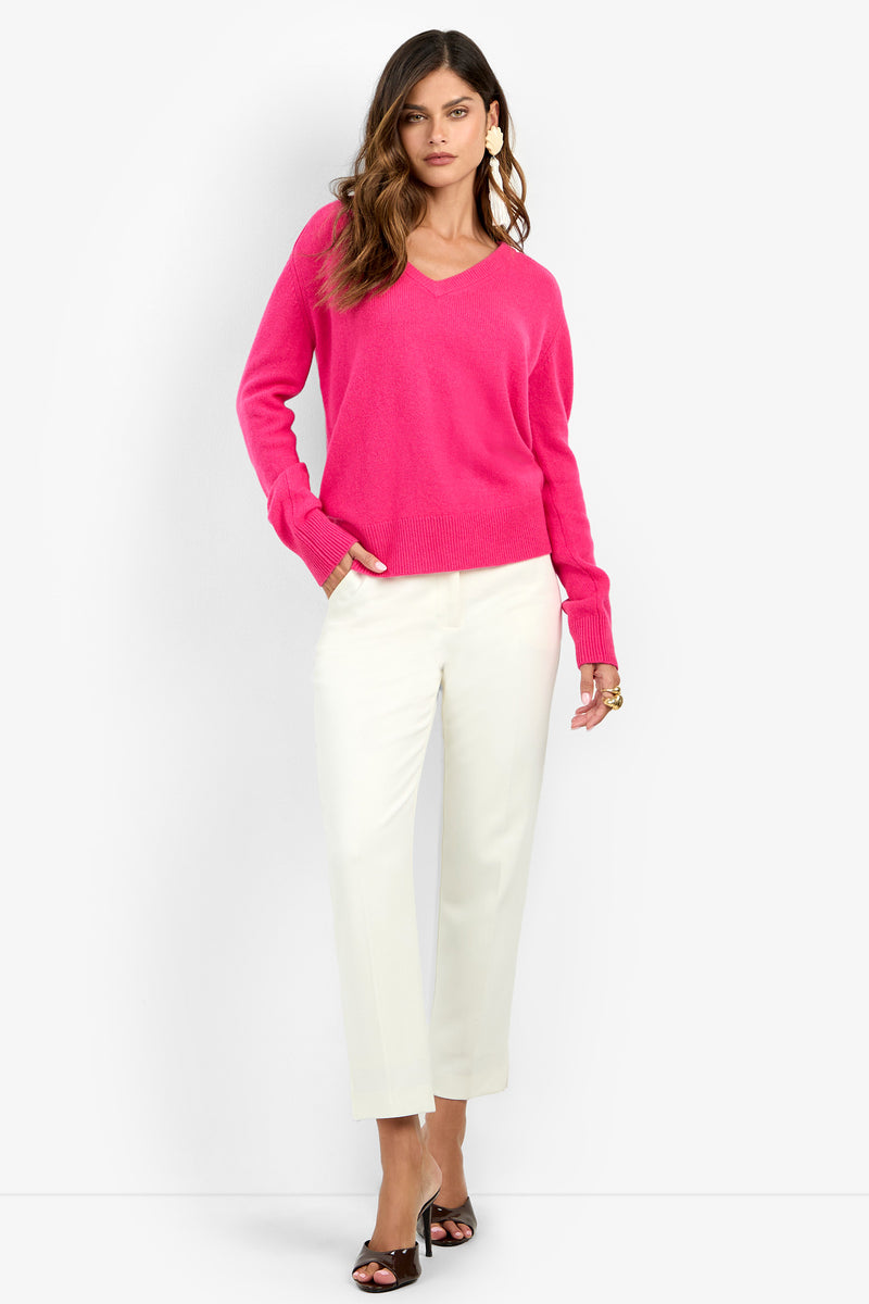 A person wears a bright pink cashmere EVANGELINE SWEATER with a V-neck, white trousers, and black heeled sandals.