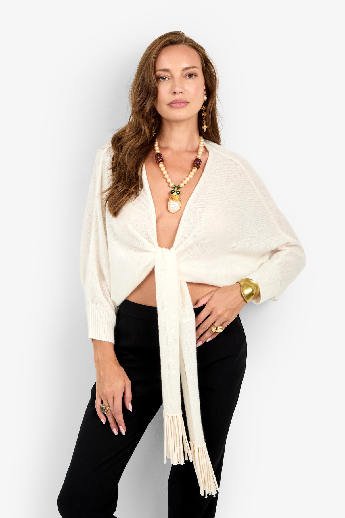 A woman wears the cream-colored knit Florentina Wrap with long sleeves, paired with black trousers and a beaded statement necklace.