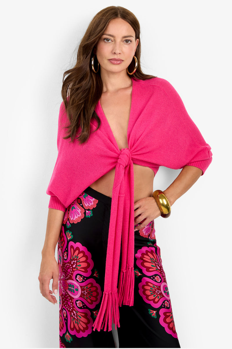 A woman wears the FLORENTINA WRAP, a bright pink cashmere sweater with a tie-front and three-quarter sleeves, paired with black pants featuring a colorful floral print.