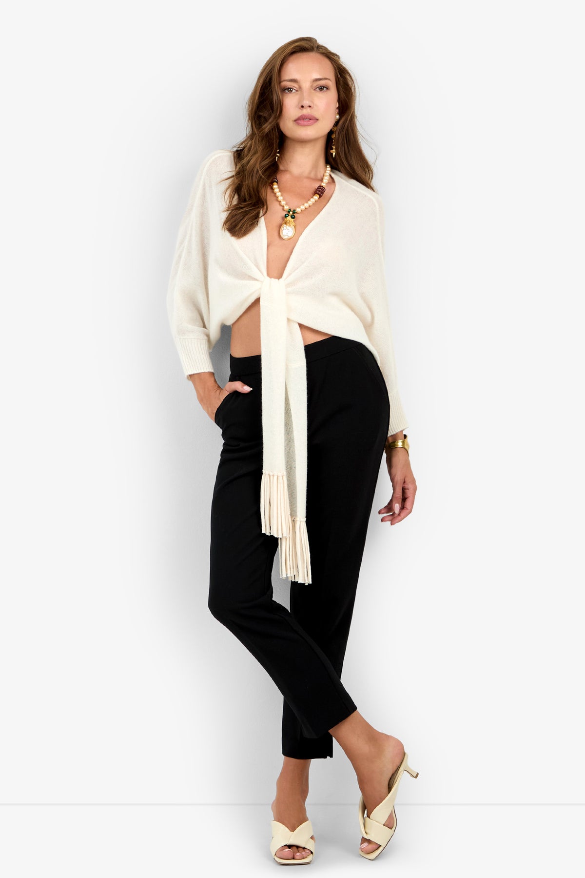 A woman poses wearing black ARNO PANT trousers, a white tie-front top, and cream-colored heeled mules.