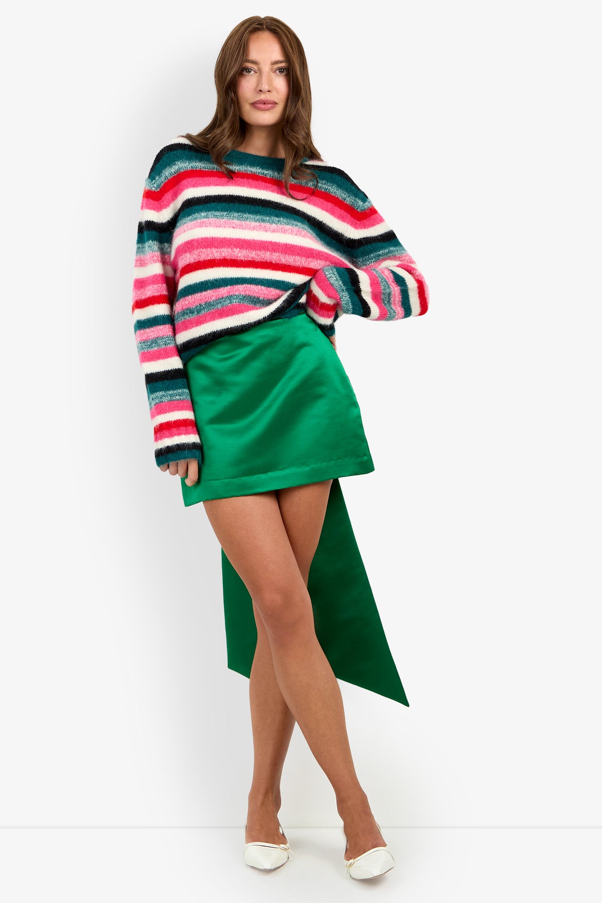 A person wears a BARDINI PULLOVER, a long-sleeved, horizontally striped sweater in pink, teal, white, and black, paired with a green mini skirt featuring a long, trailing back panel, white pointed-toe heels, and nylon socks.