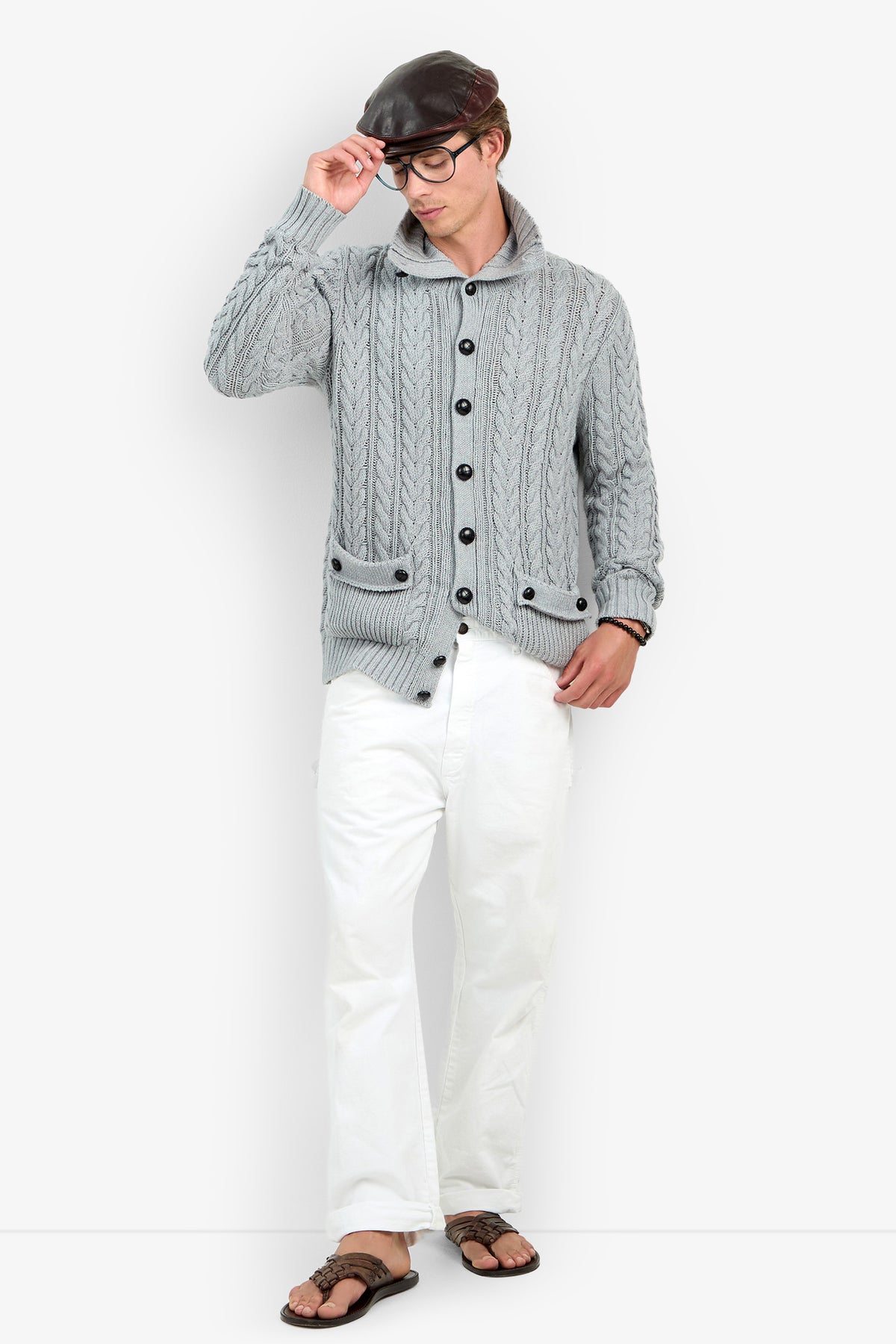 A person wearing the RALEIGH SWEATER COAT in HEATHER GREY, paired with white pants, black glasses, and a black cap, stands while touching the brim of the cap with one hand and looking down.