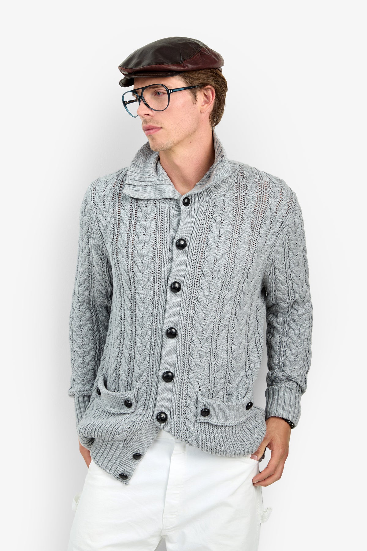 A person wearing the RALEIGH SWEATER COAT, styled with white pants, glasses, and a dark flat cap, stands against a white background with hands in pockets, looking to the side.