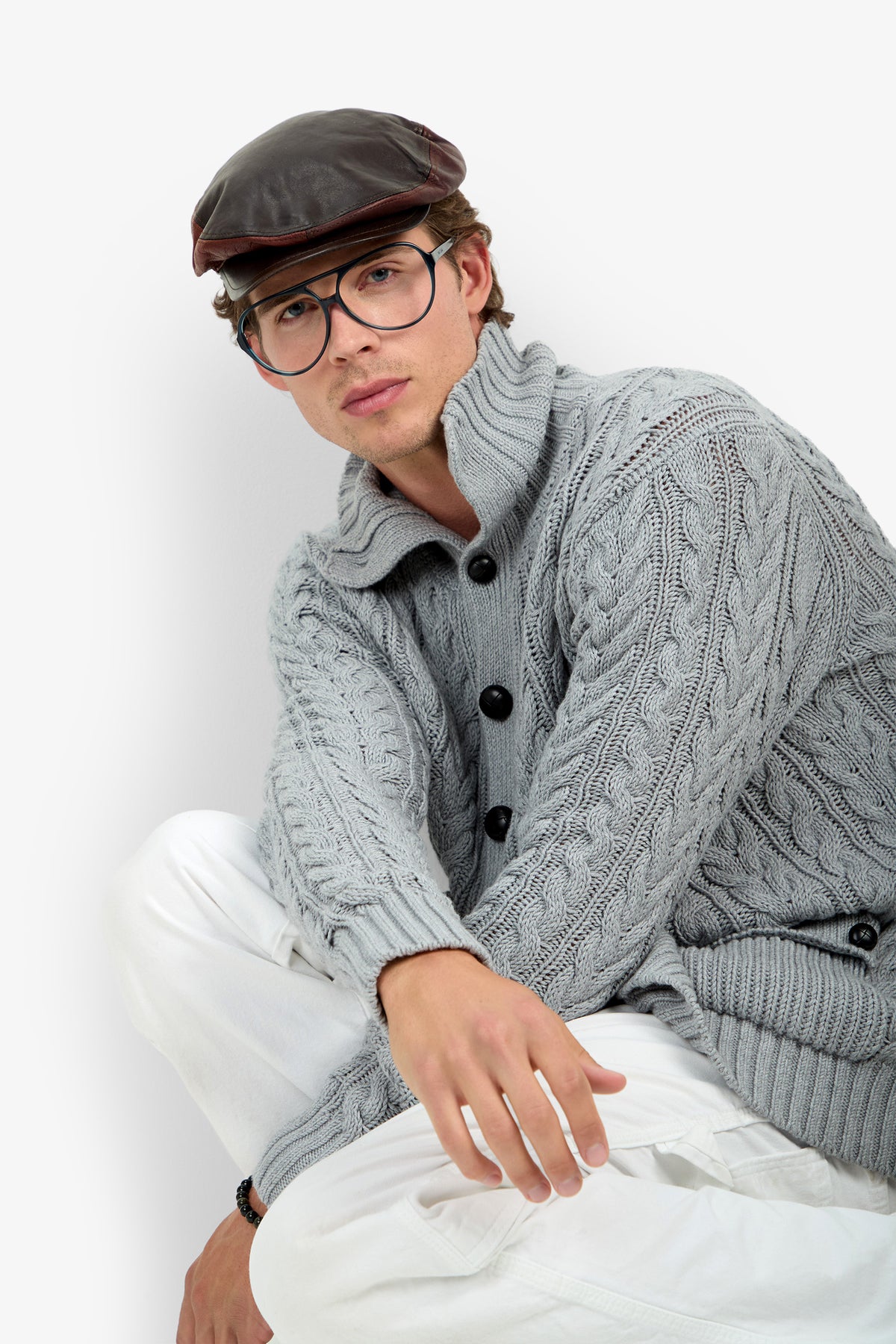 A person wearing the RALEIGH SWEATER COAT, styled with white pants, large glasses, and a brown leather cap, sits against a white background.