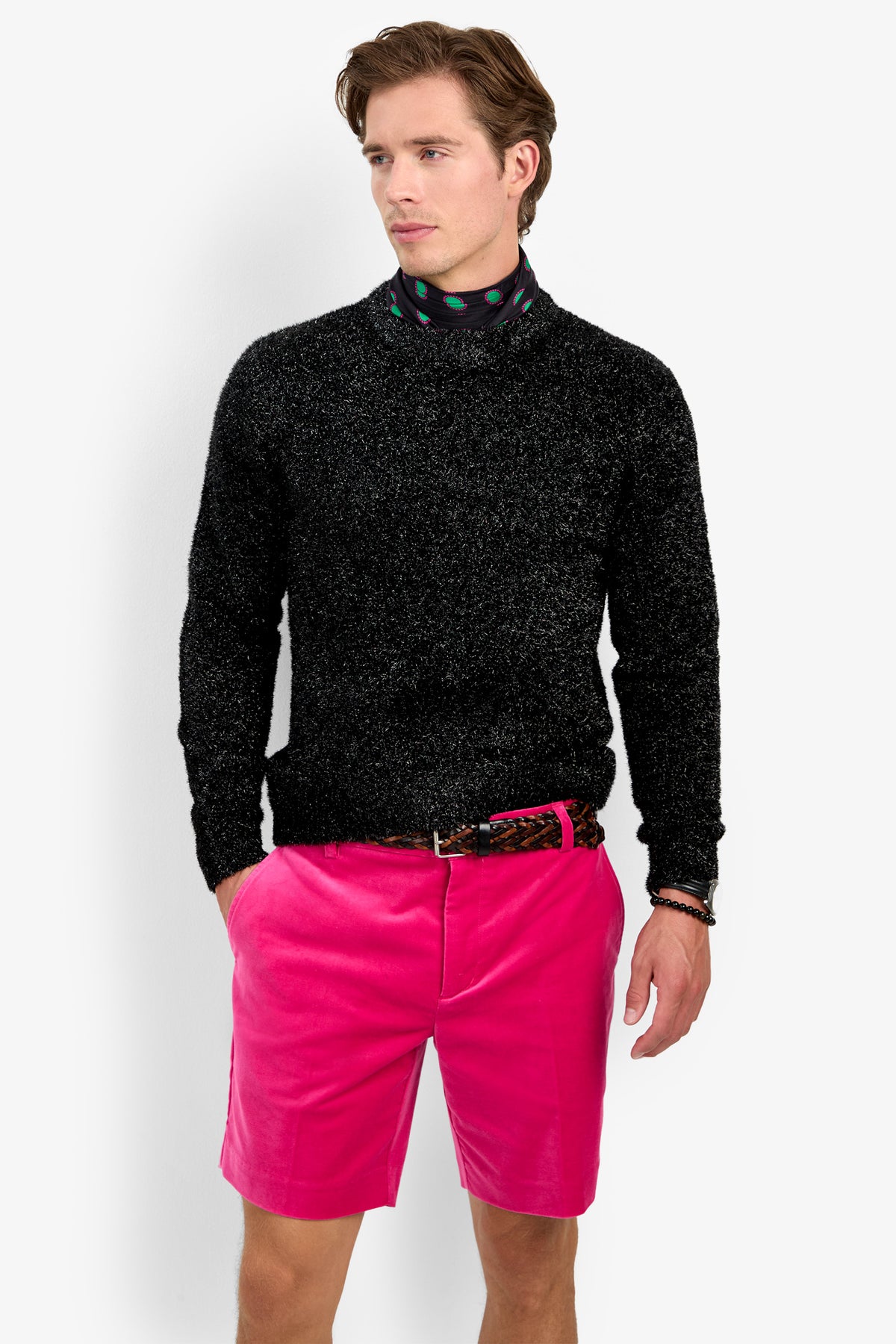 A man wearing the ASTRON SPARKLE CREWNECK, a black scarf with colorful dots, bright pink shorts, and a patterned belt stands with his hands in his pockets, looking to the side.