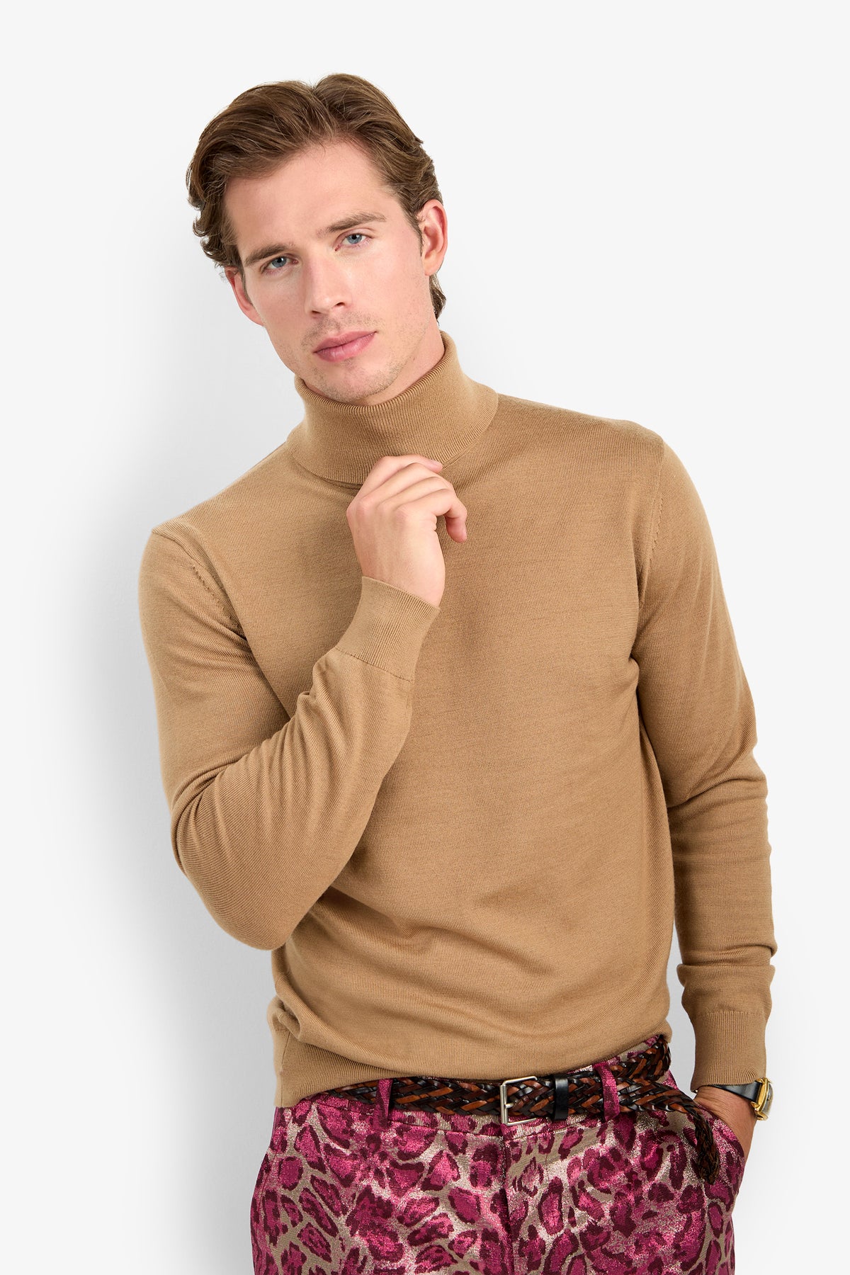 Wearing the WOODROW TURTLENECK in tan and pink leopard-print pants, a man stands against a white background, gazing at the camera with a relaxed expression.