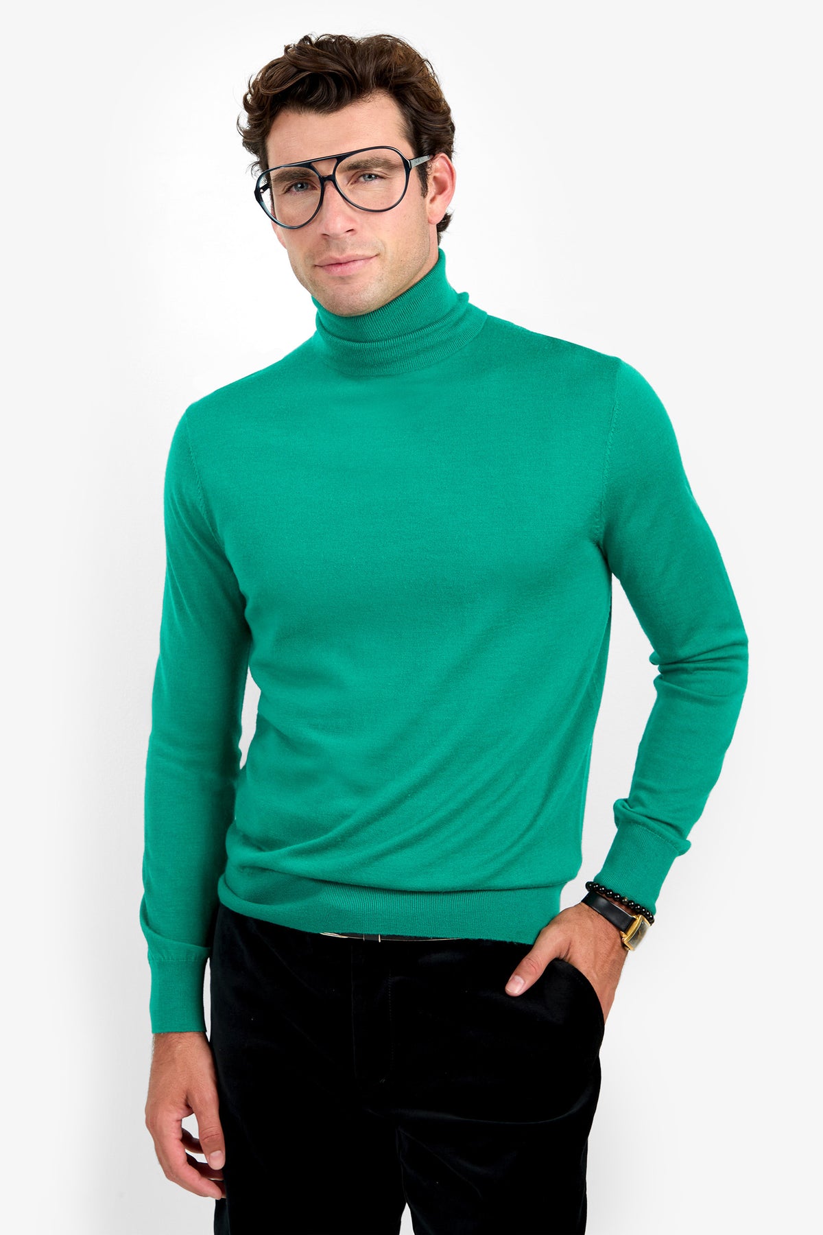 A man wearing large black glasses and the WOODROW TURTLENECK in green stands with one hand in his pocket, looking at the camera against a plain white background.