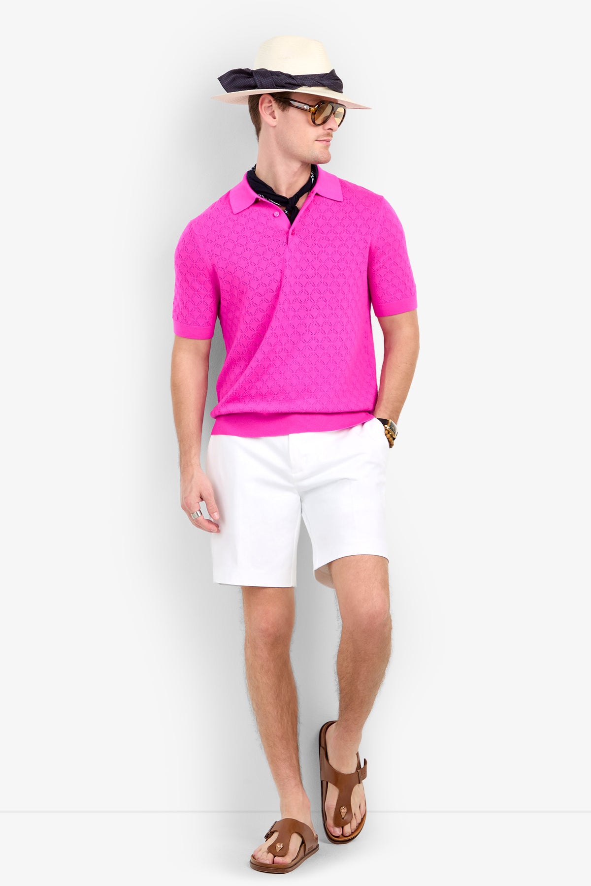 A man wearing a textured bright pink Waylen Polo, white shorts, a straw fedora, sunglasses, and leather sandals.