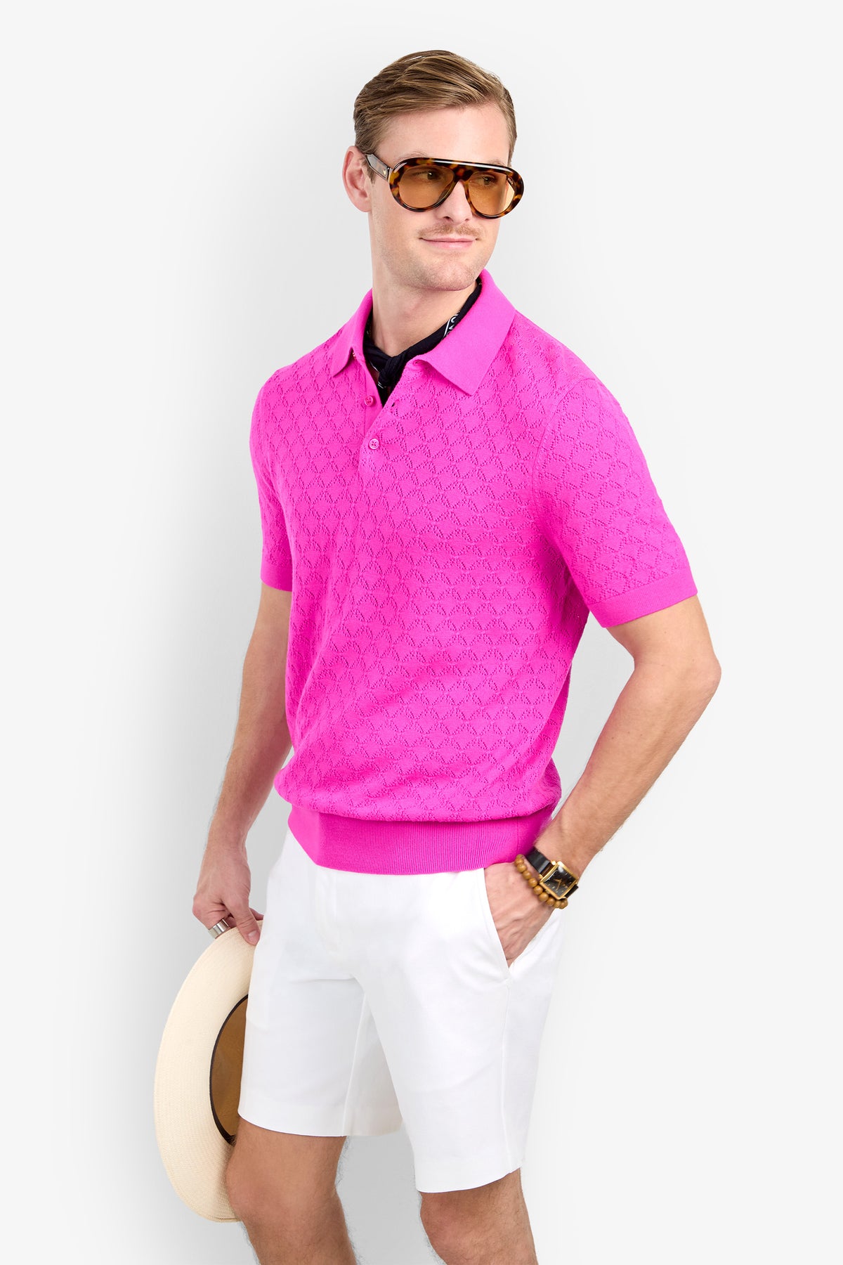 A man wearing a textured, short-sleeved pink Waylen Polo, white shorts, and oversized sunglasses, holding a straw hat.