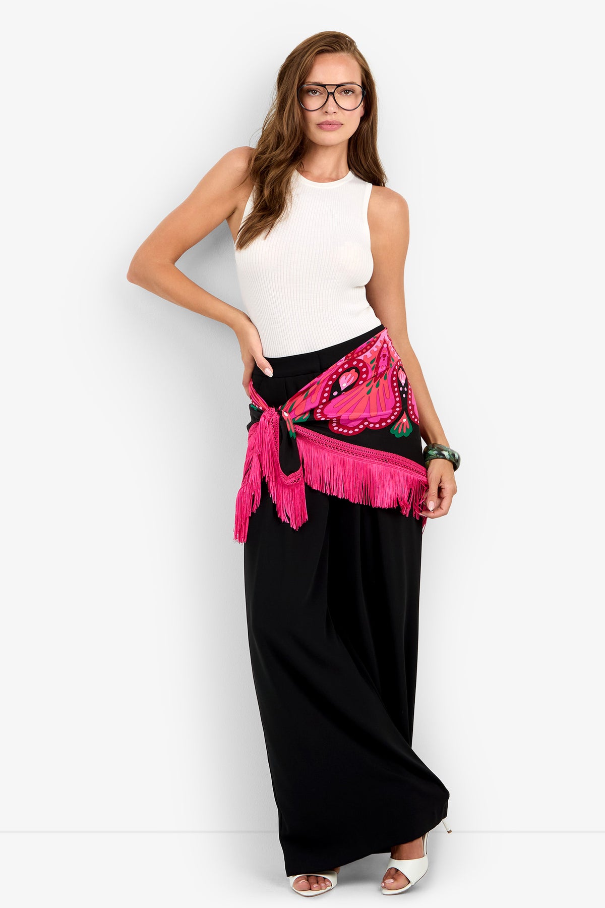 Woman stands against a white background wearing glasses, a white sleeveless top, long black skirt, white heels, and the Sautoir Fringe Scarf in bright pink tied at her waist.
