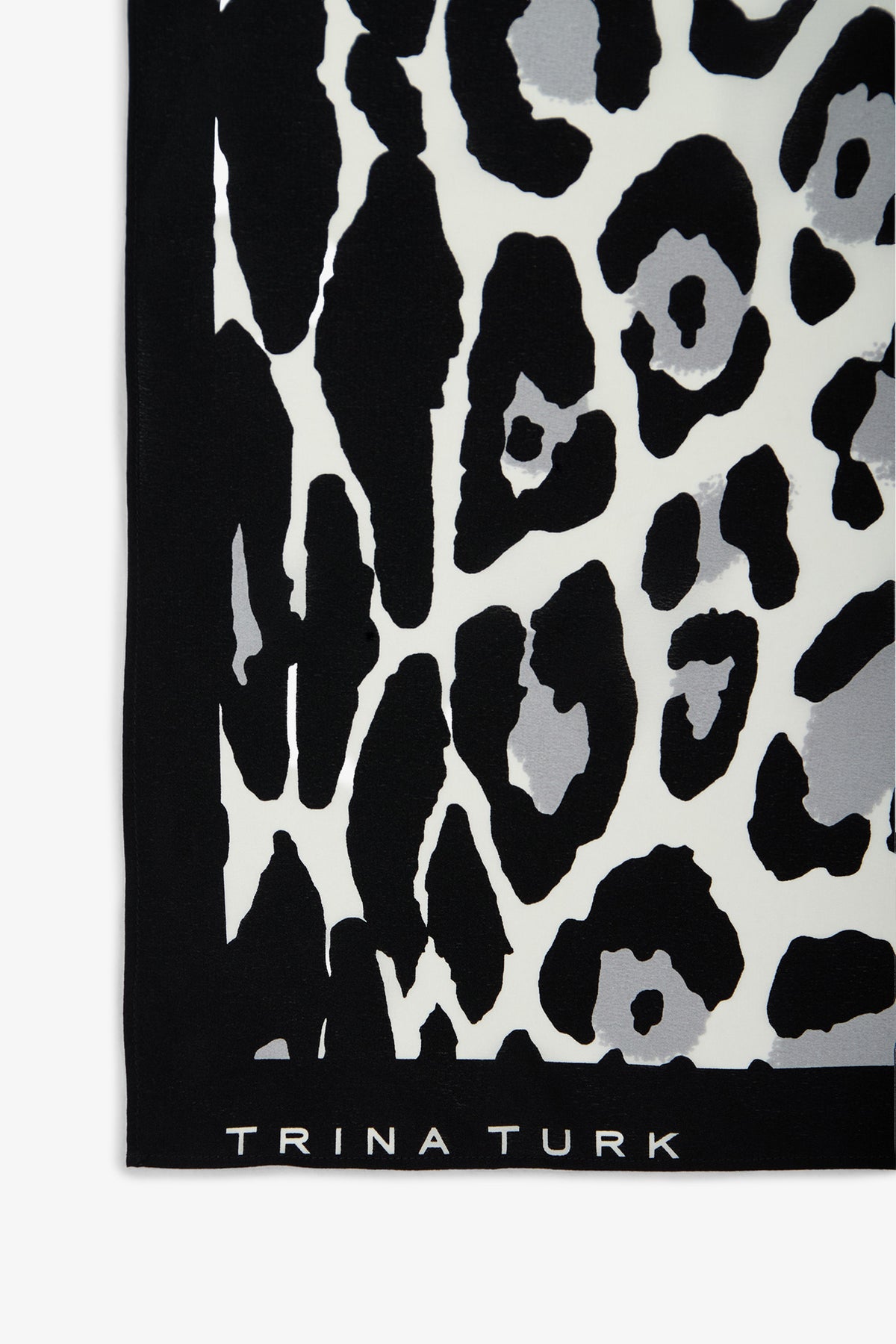 A section of the Snow Leopard Silk Scarf featuring a black, white, and gray leopard print pattern, with “TRINA TURK” displayed in white along the bottom edge.