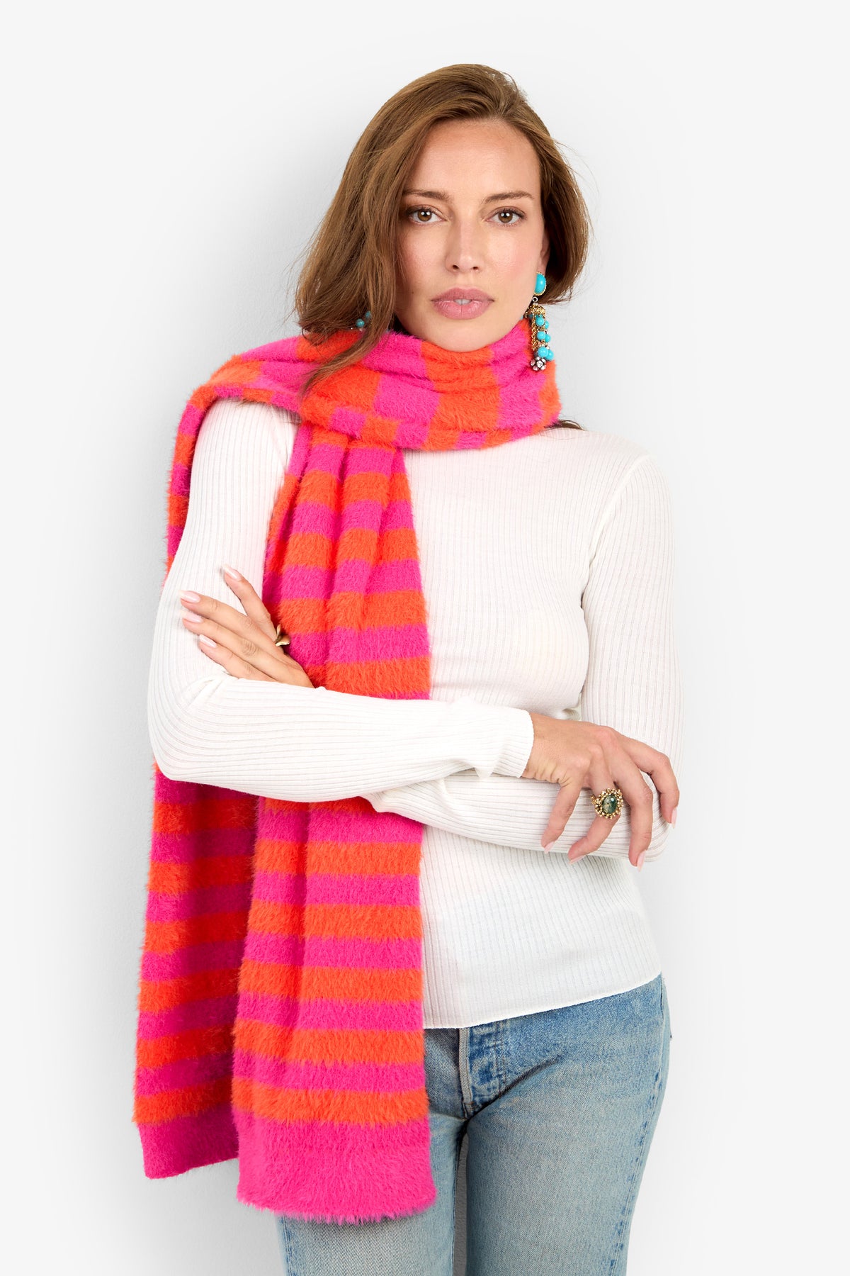 A woman in a white ribbed sweater and blue jeans wears the RAJA SCARF, a pink and orange striped scarf. She has long brown hair, blue earrings, and stands with arms crossed against a plain white background.