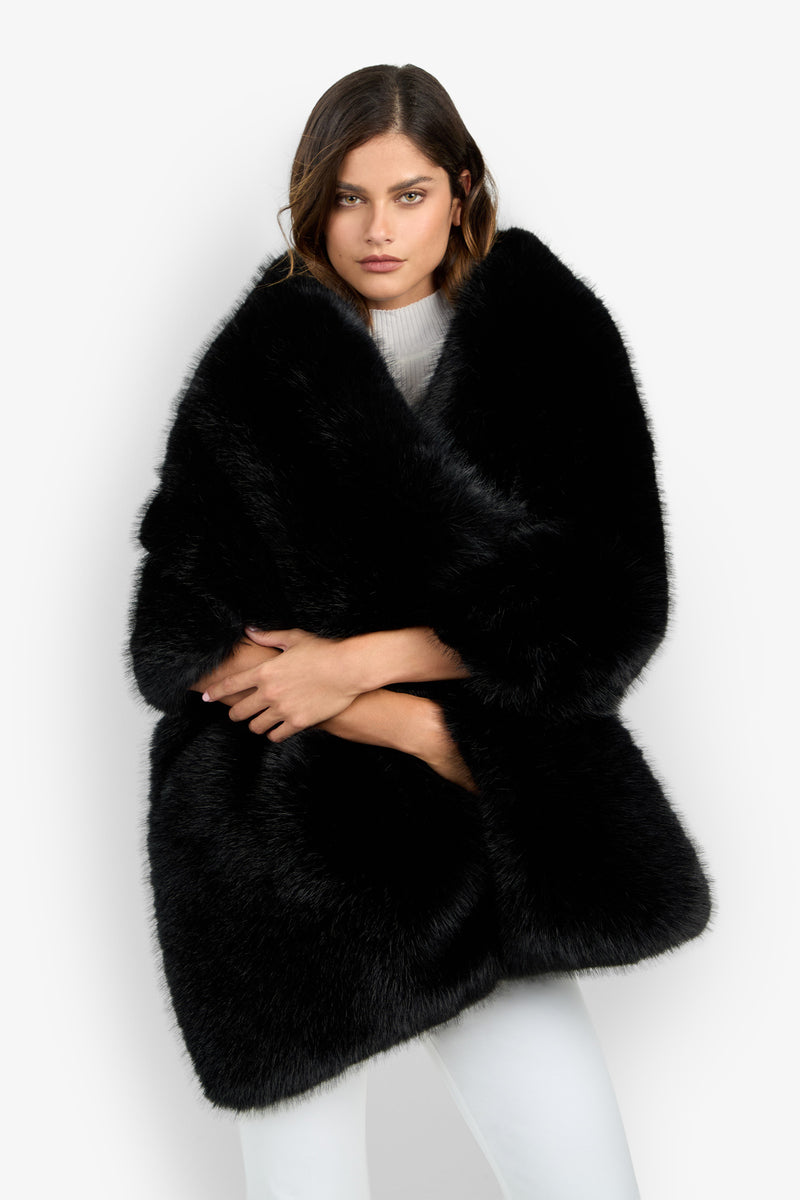 A woman stands against a plain white background with her arms crossed, wearing the Pellicia Fur Stole in BLACK over a white top and white pants.