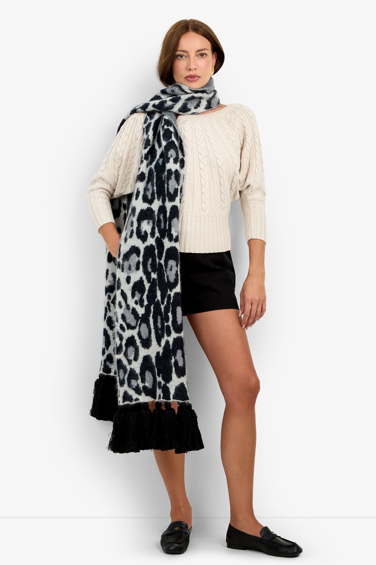 A woman wears a CARDUCCI LEOPARD SCARF in long, black and grey wool cashmere with black fringe ends over a cream-colored sweater and black shorts.