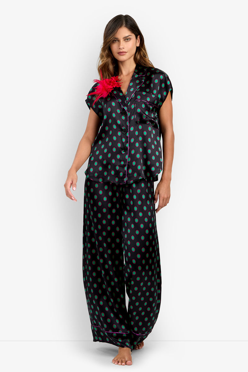 A person wearing the ADARA 2 TOP, a 100% silk button-up pajama set with a black and teal polka dot pattern and a bright pink feather accent on the collar.