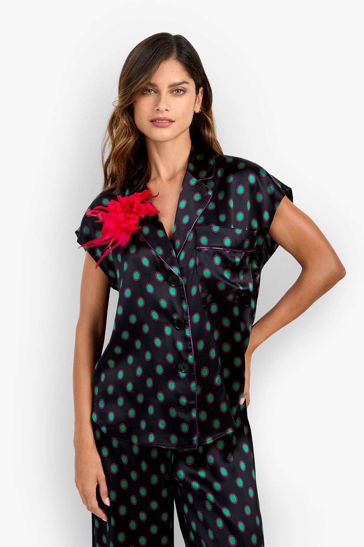 A woman wears the ADARA 2 TOP, a black, short-sleeved 100% silk button-down with a green and magenta circular pattern and a bright pink feathered flower pinned to the chest.