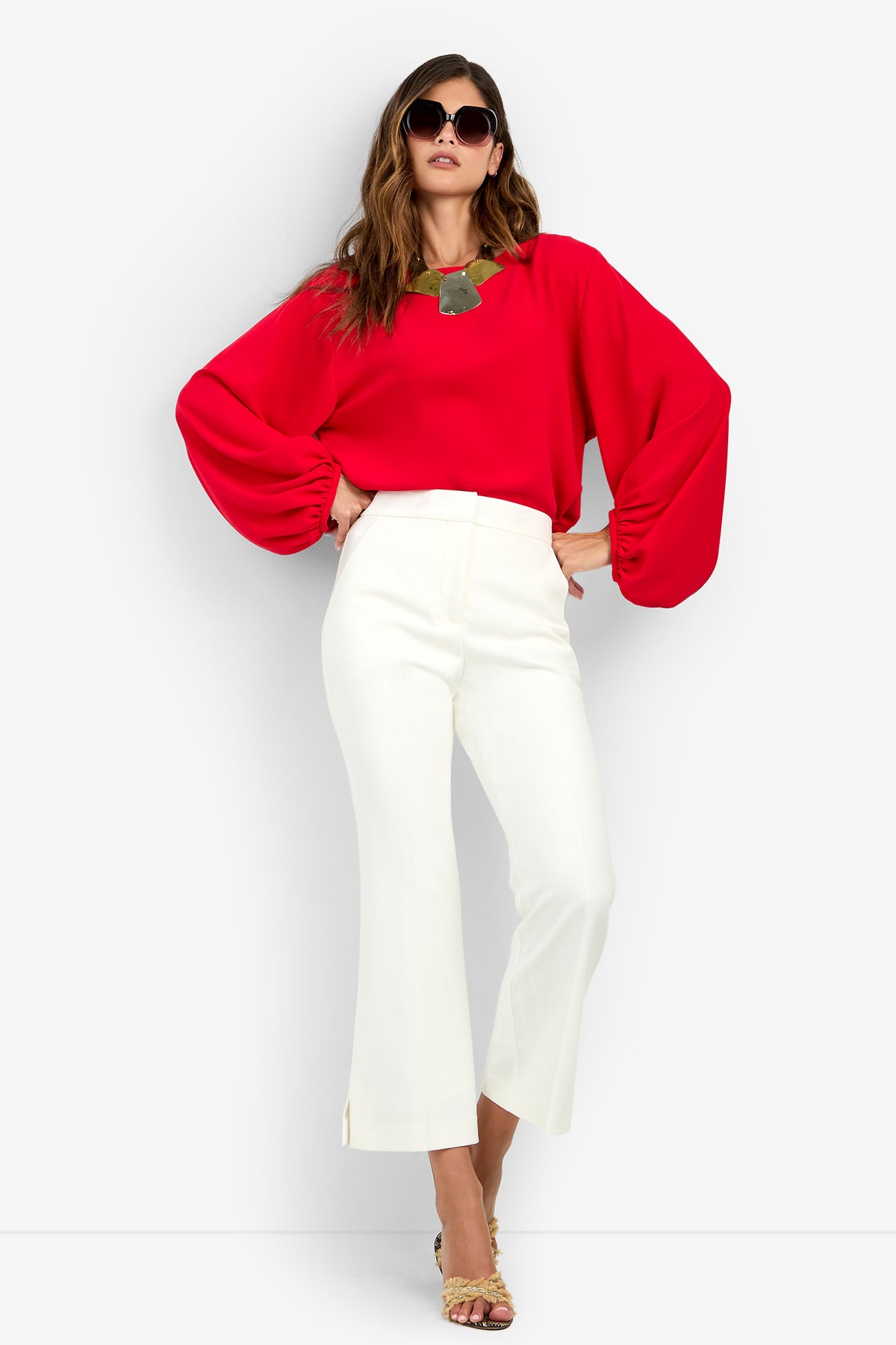 A woman wears the URSA 2 TOP, a 100% polyester red blouse with billowy long sleeves, paired with white cropped trousers and sunglasses.