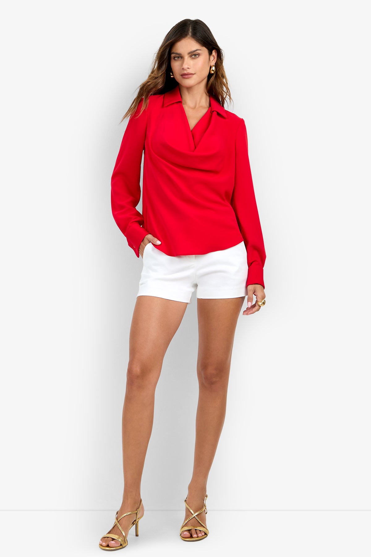 A woman wears a MISTIC TOP, a long-sleeved red cowl-neck piece in polyester fabric, paired with white shorts and gold strappy heels.