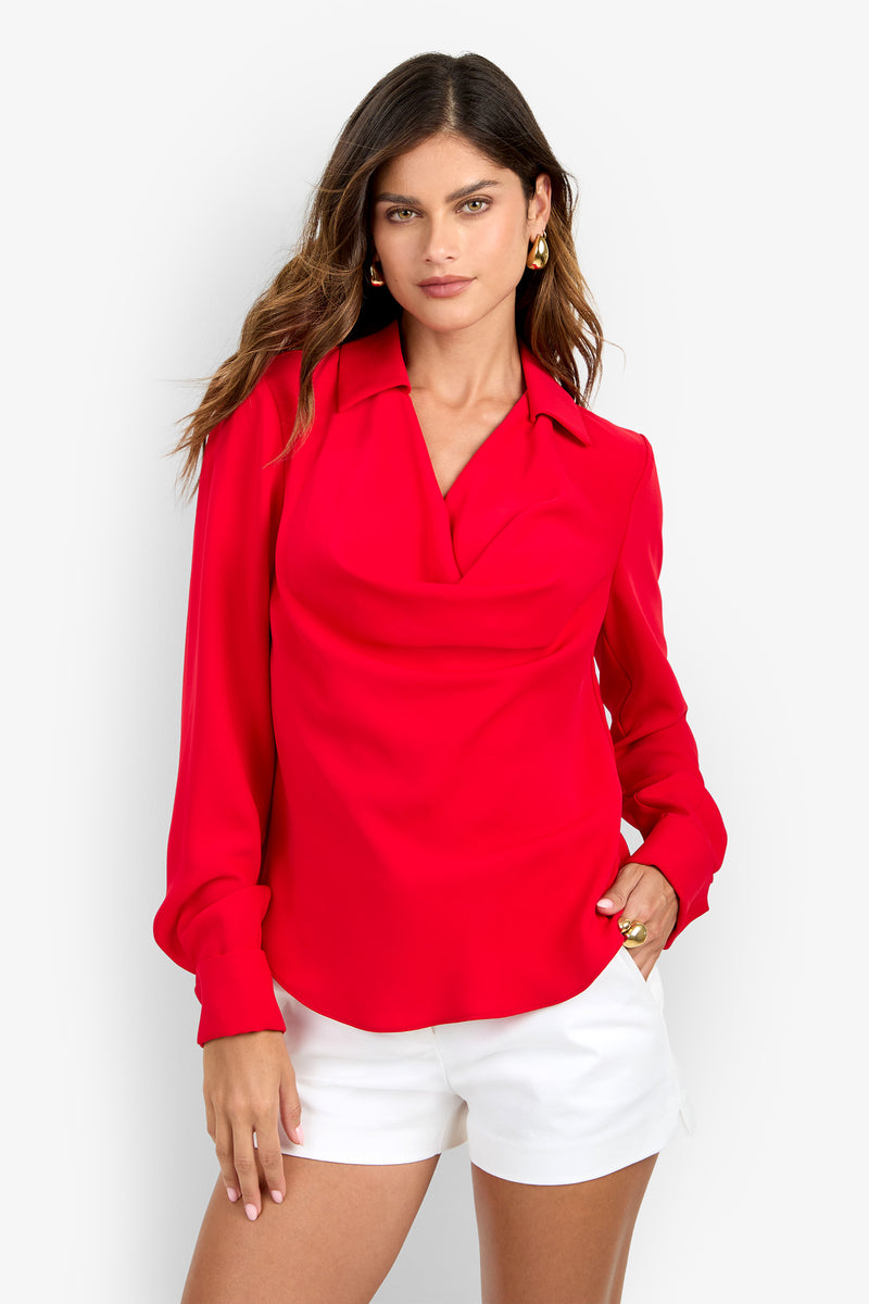 A woman wears a long-sleeved, bright red polyester MISTIC TOP with a draped cowl neckline and white shorts.