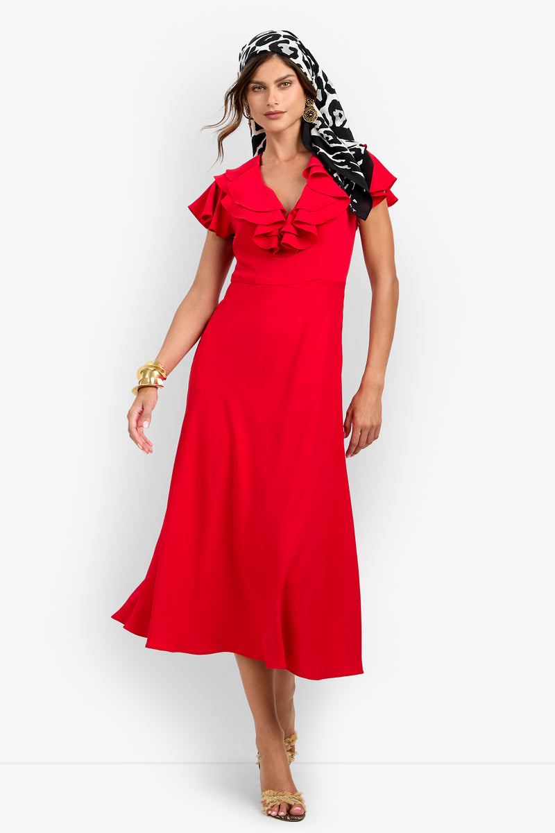 A woman wears the red polyester PATRONS DRESS featuring ruffled sleeves and a ruffled V-neckline, styled with a black-and-white patterned headscarf, gold cuffs, and metallic heels.