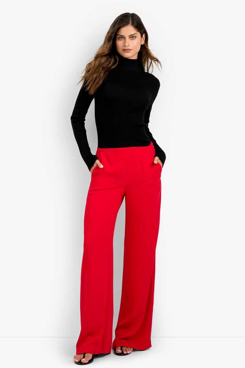 A woman wears a black turtleneck and TAVIA PANT, featuring bright red wide-leg trousers made of 100% polyester.