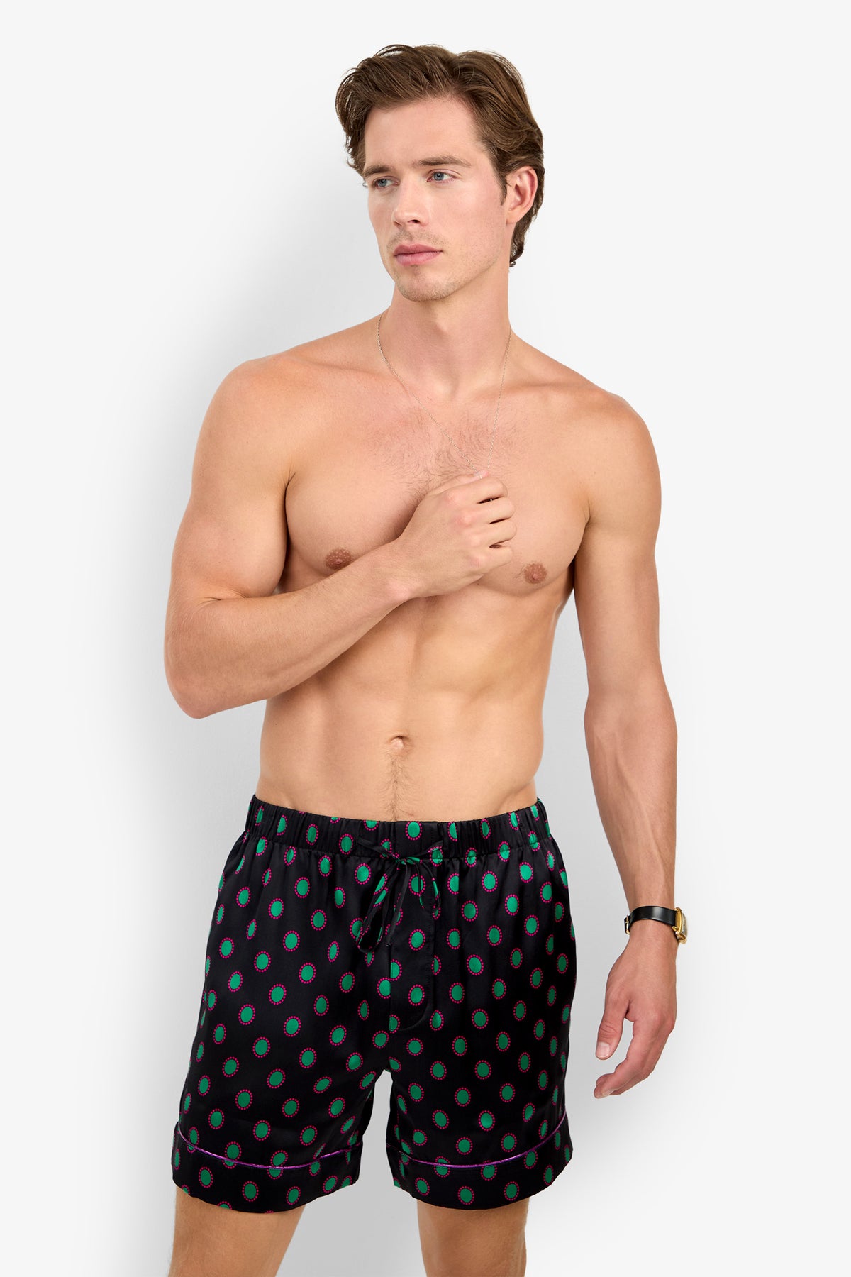 A shirtless man wearing PJ SHORT black pajama shorts made of pure silk with a repeating teal and magenta circular pattern.