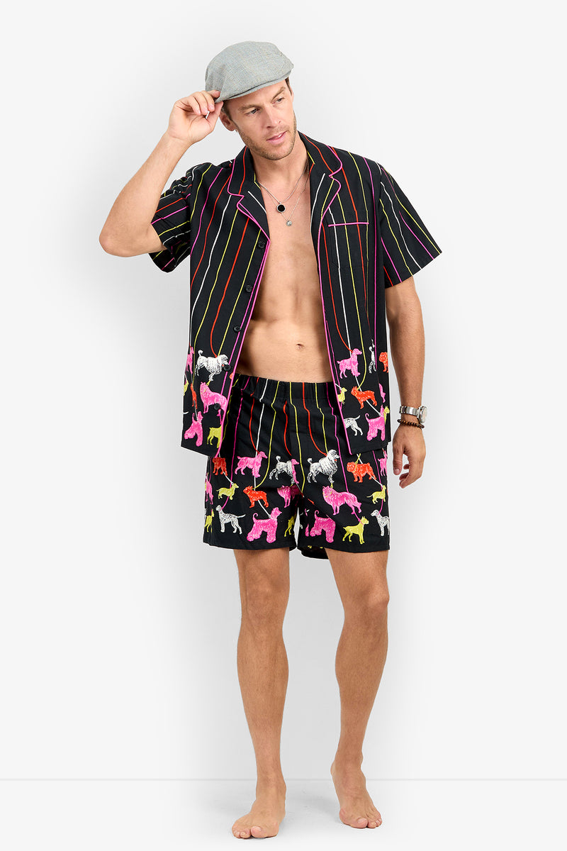 A man wears a PJ SHORT set consisting of a short-sleeved, button-down shirt and matching shorts in a black fabric featuring vertical stripes and colorful dog silhouettes.