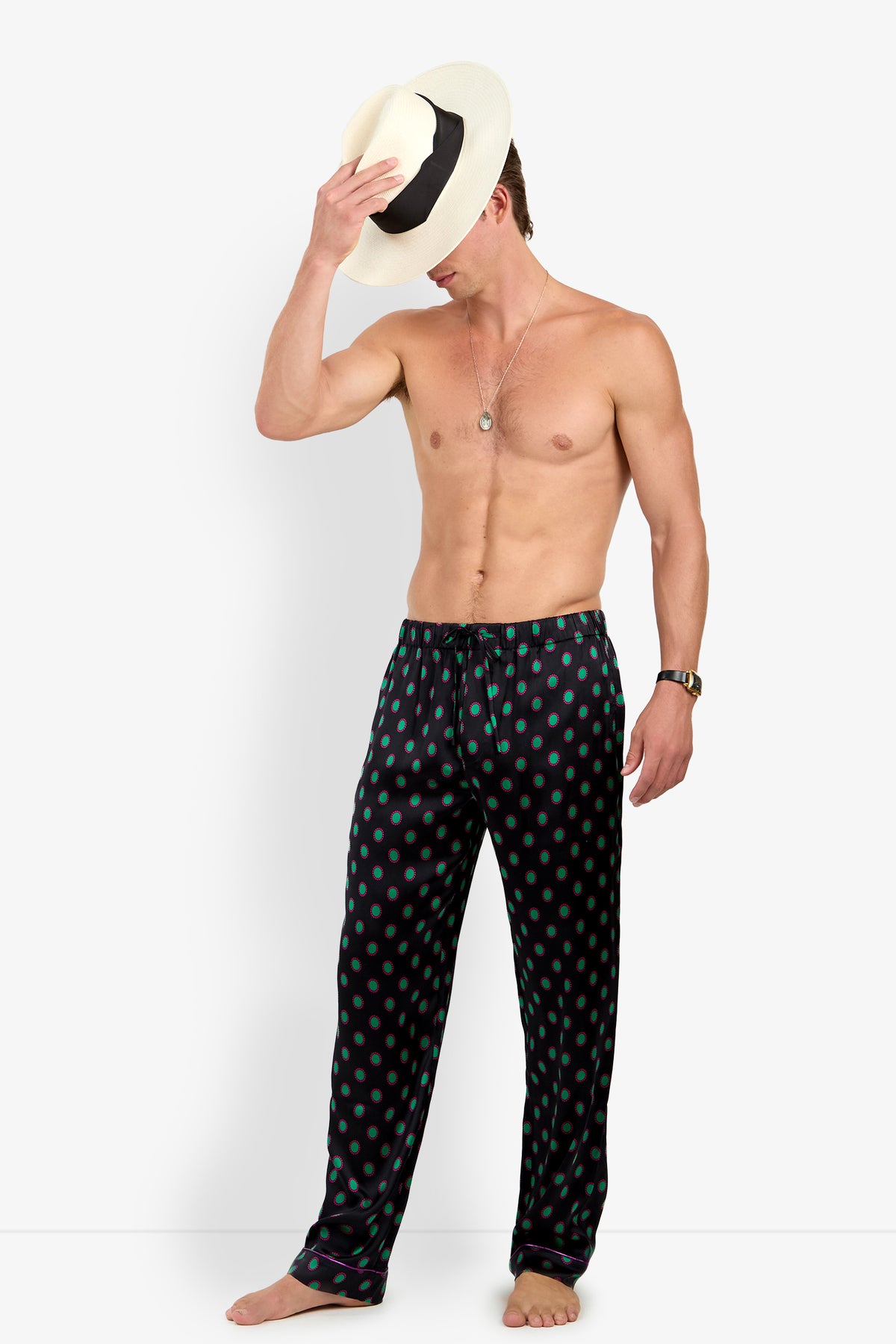 A shirtless man wears PJ PANT in black pure silk with a green and pink geometric print while holding a straw fedora.