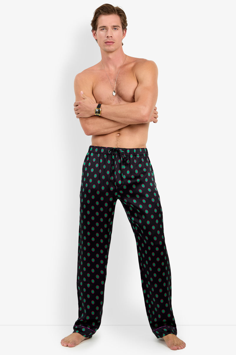 A shirtless man wearing black PJ PANT silk pajama pants featuring a repeating green and pink circular pattern.
