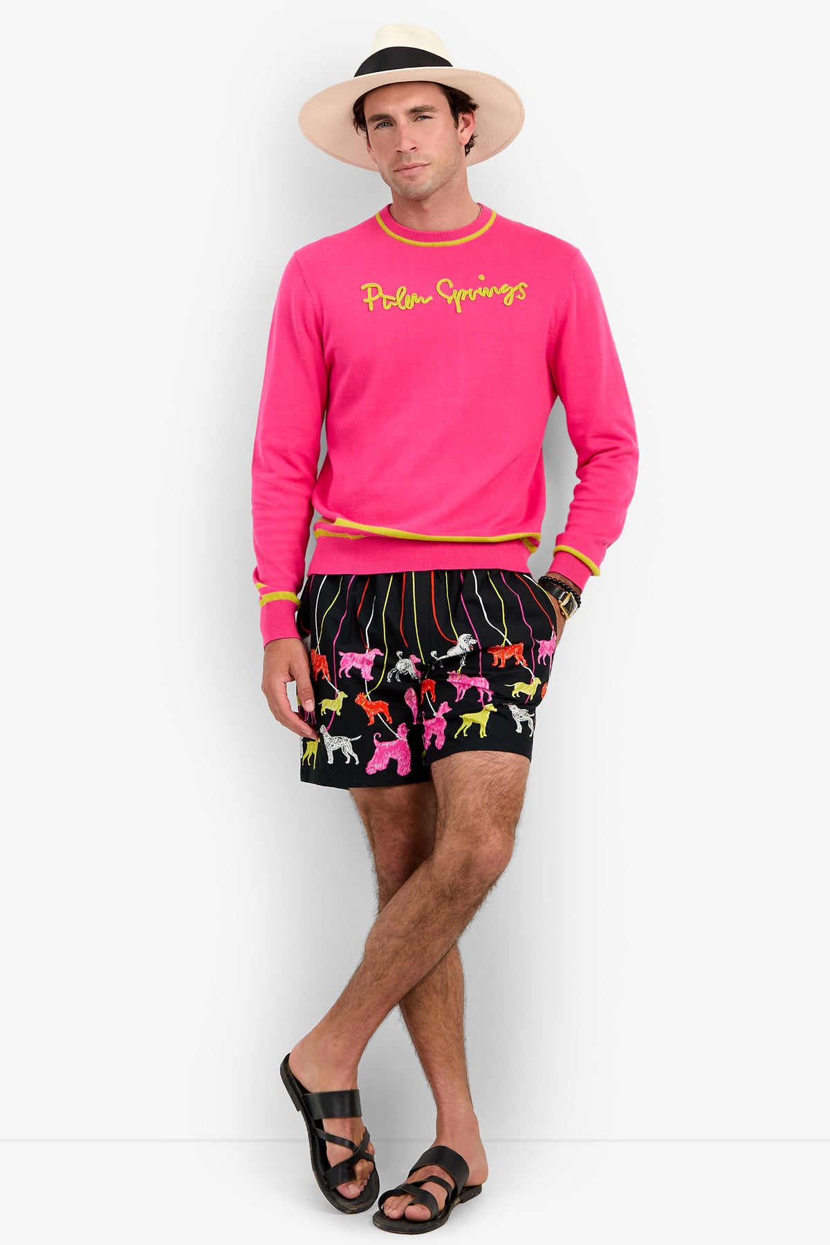 A man wears a bright pink PALM SPRINGS CREWNECK with yellow trim and embroidered chest text, paired with black shorts featuring a colorful dog print and black leather sandals.