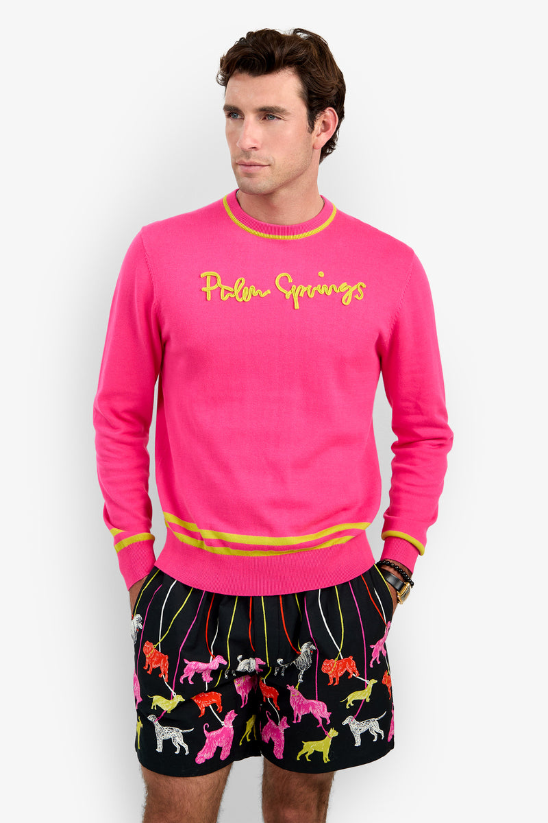 A man wears a bright pink cotton modal PALM SPRINGS CREWNECK with yellow embroidery and striped trim, paired with black shorts featuring a colorful dog print.
