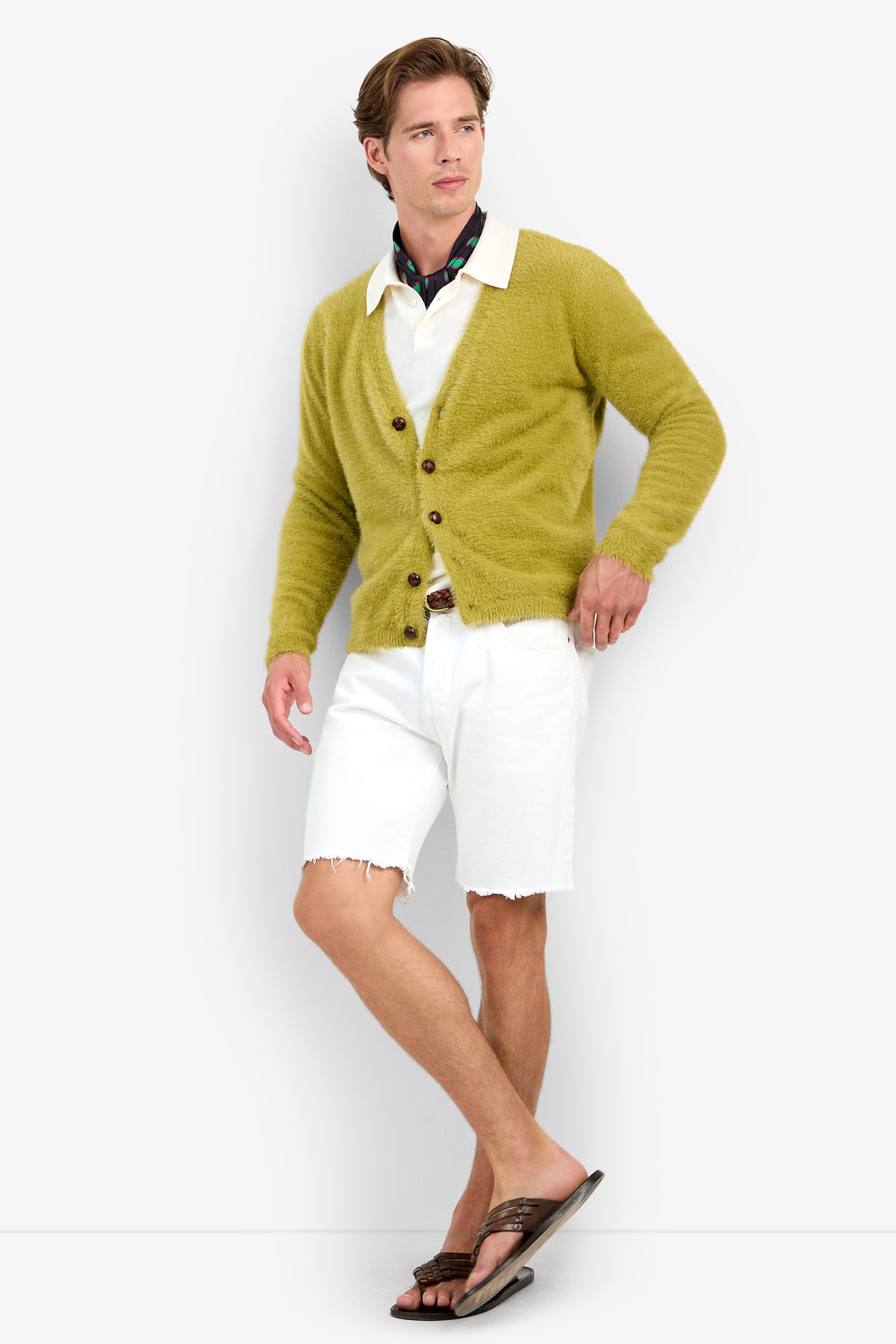 A man wears a fuzzy olive-green SCOTT FUZZY CARDIGAN over a white collared shirt and white denim shorts with frayed hems, finished with a patterned neck scarf and leather flip-flops.