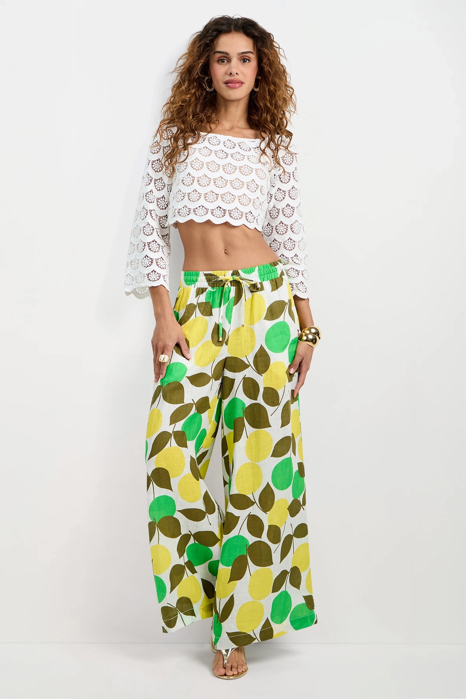 A woman stands against a plain white background wearing the Azule Wide-Leg Pants with a green, yellow, and brown leaf pattern, paired with a white crochet crop top. She has long curly hair and accessorizes with bangles and rings.