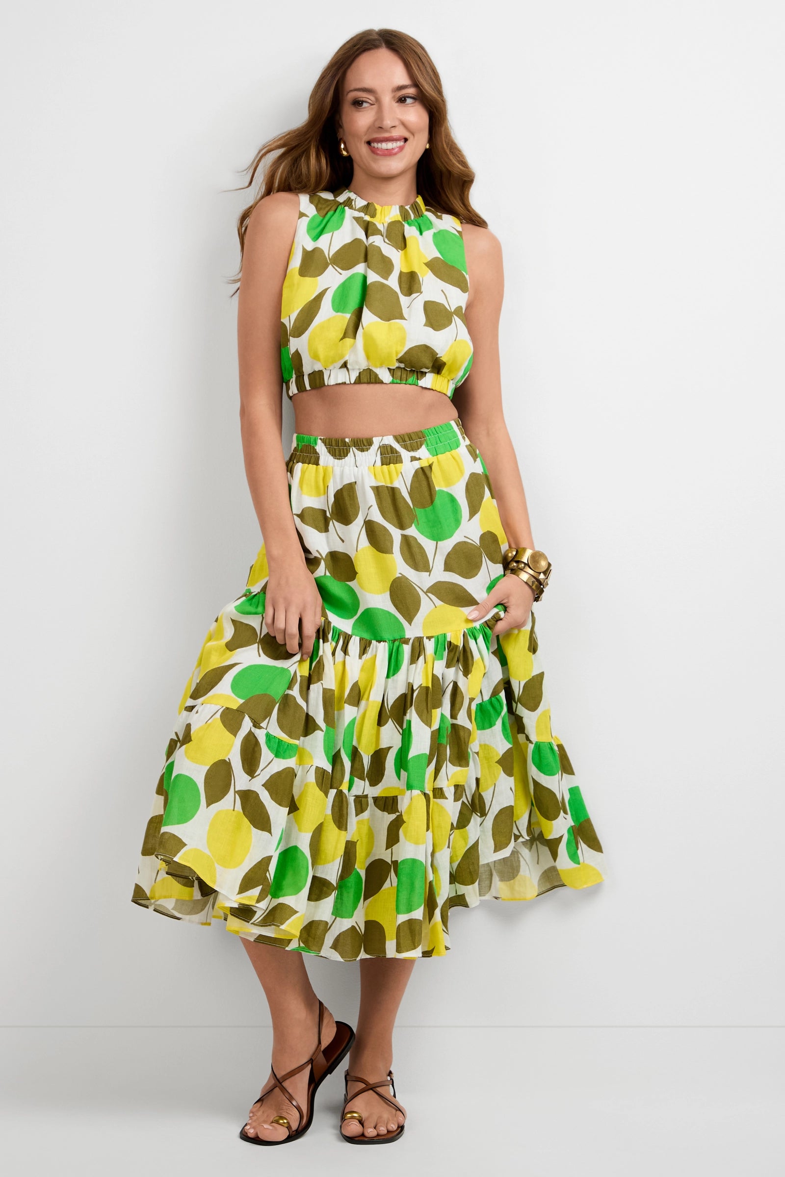 A woman smiles while wearing the Marsay Drop Waist Midi Skirt and a matching sleeveless, high-neck crop top in a bold green, yellow, and brown leaf print. She accessorizes with bracelets and brown sandals against a plain white background.
