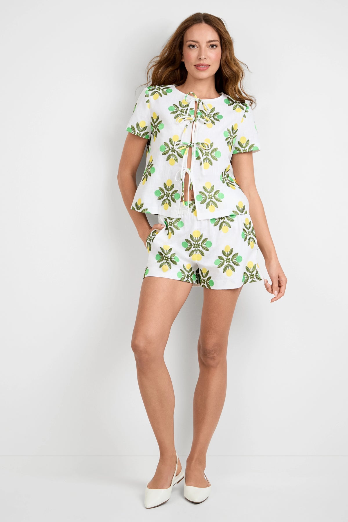 A woman with long, wavy hair stands against a white wall wearing Azule Shorts and a matching white short-sleeve top with green and yellow floral patterns, paired with white pointed-toe shoes.