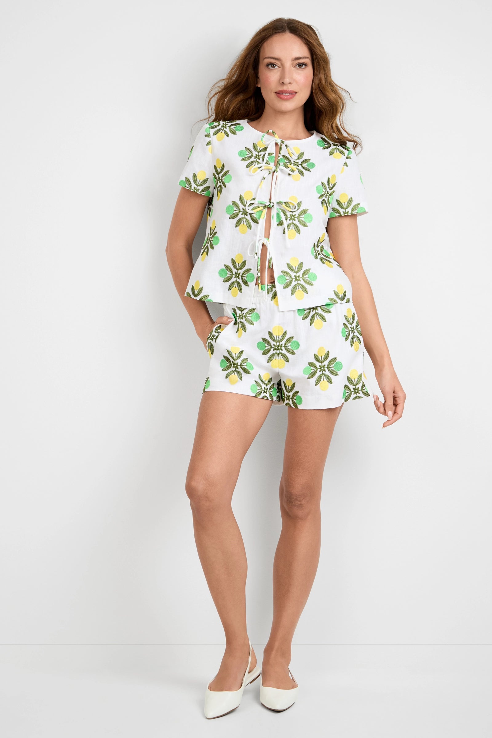 A woman with long, wavy hair stands against a white wall wearing Azule Shorts and a matching white short-sleeve top with green and yellow floral patterns, paired with white pointed-toe shoes.