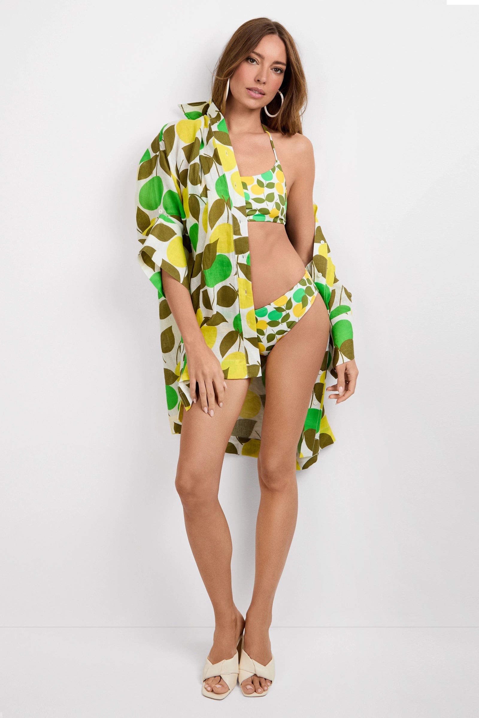 A woman with long brown hair and hoop earrings poses in the Arissa Hipster Swim Bottom - Limone, a green, yellow, and white leaf-patterned bikini set with a matching open shirt and beige heeled sandals against a white background.