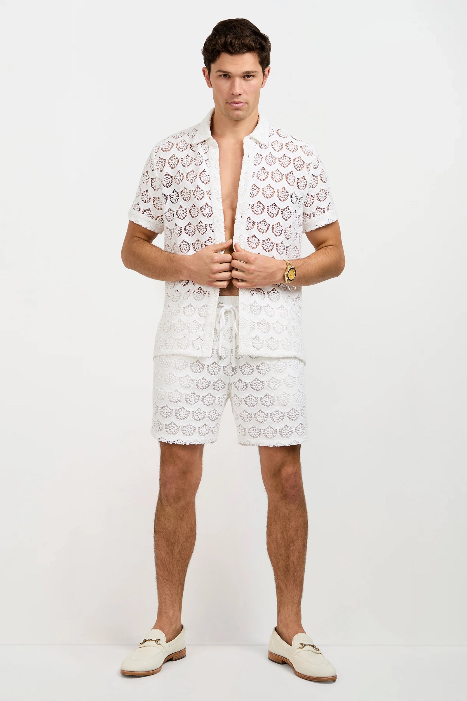 A man faces forward wearing the Branson Crochet Lace Shirt—white, short-sleeved, button-up with a lacy pattern—paired with matching shorts, white loafers, and a gold wristwatch. The background is plain white.