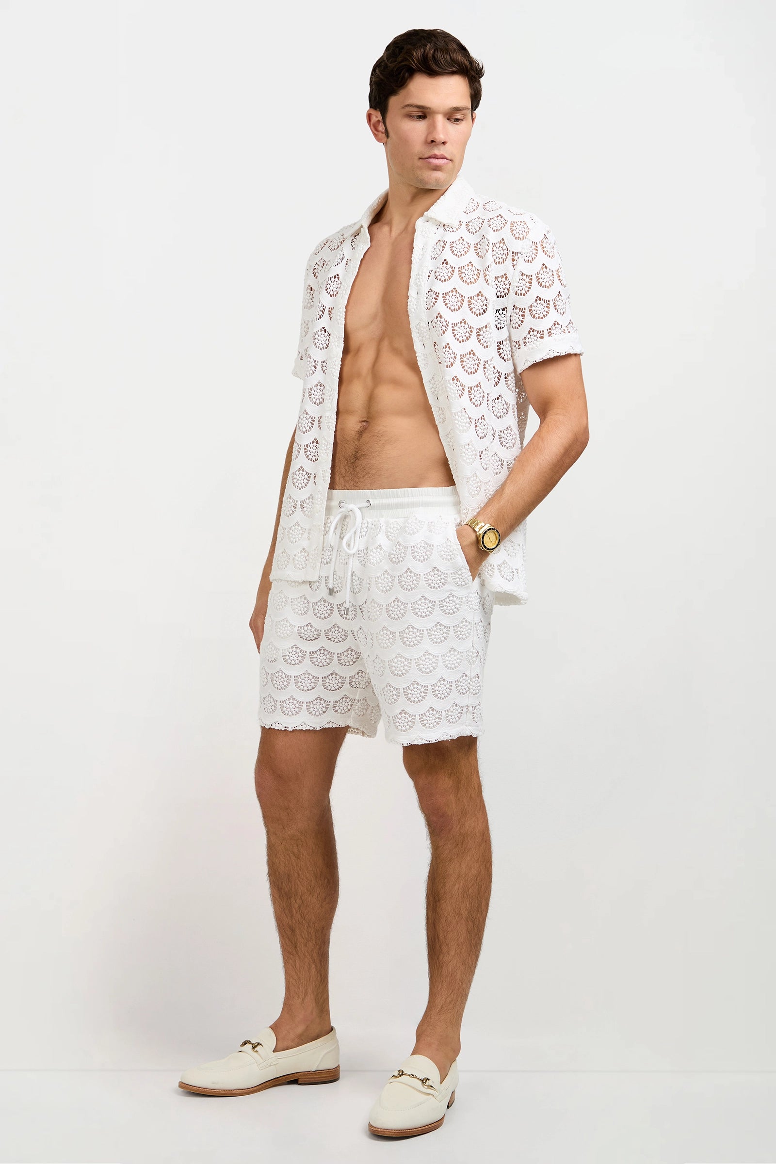 A man stands against a plain background wearing the Branson Crochet Lace Shirt, unbuttoned to reveal his chest, paired with matching drawstring shorts, cream loafers, and a watch.