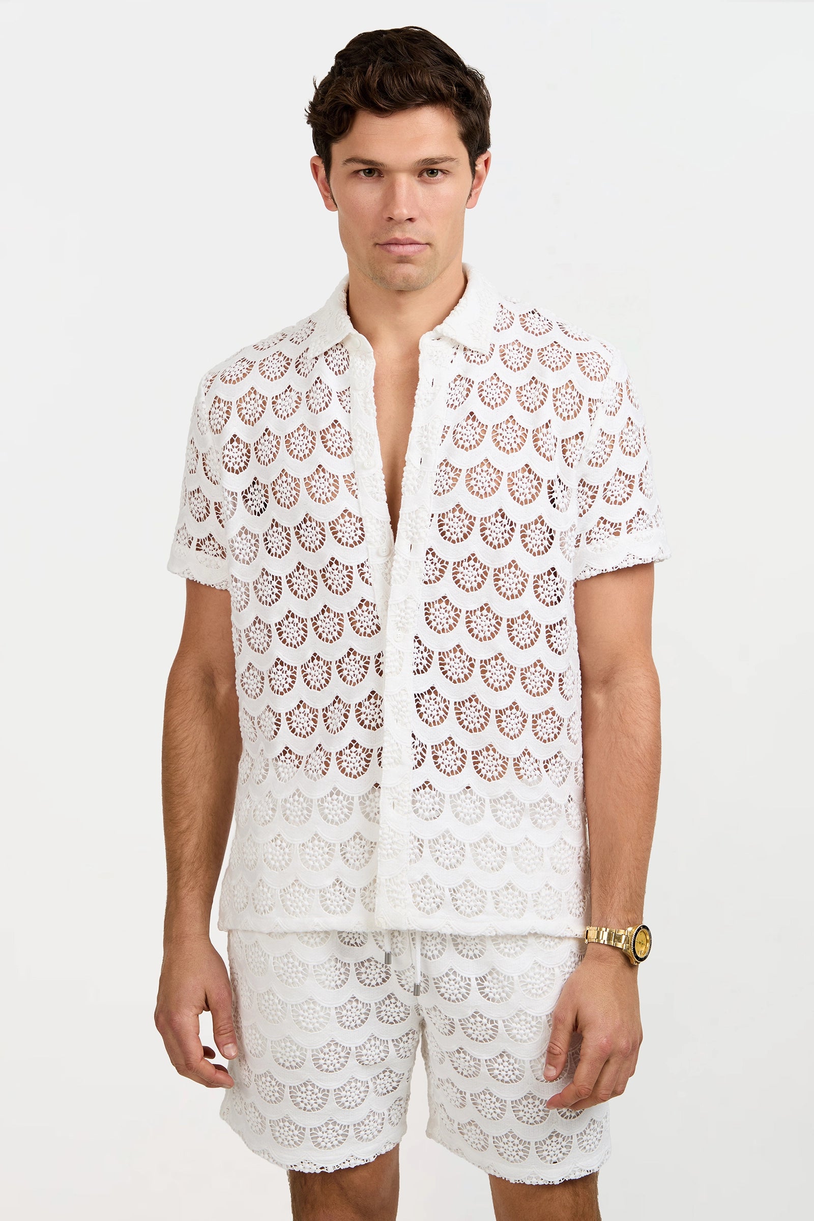 A man with short brown hair and a neutral expression stands against a plain light background wearing the Branson Crochet Lace Shirt, matching white see-through shorts, and a gold wristwatch.