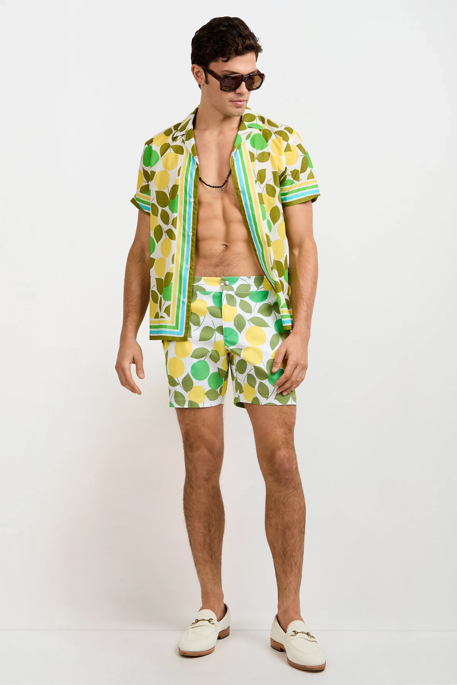 A man wearing sunglasses, the Josue Short-Sleeve Shirt - Limone, matching leaf-print shorts, white loafers, and a beaded necklace stands against a plain white background.