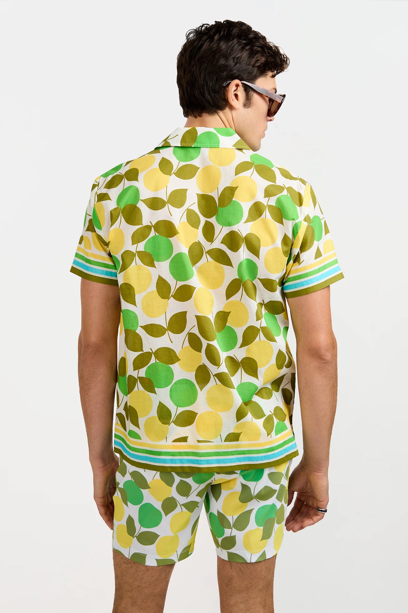 A man wearing the Josue Short-Sleeve Shirt - Limone, featuring a bold green and yellow leaf pattern, stands with matching shorts and sunglasses, facing away against a white background.