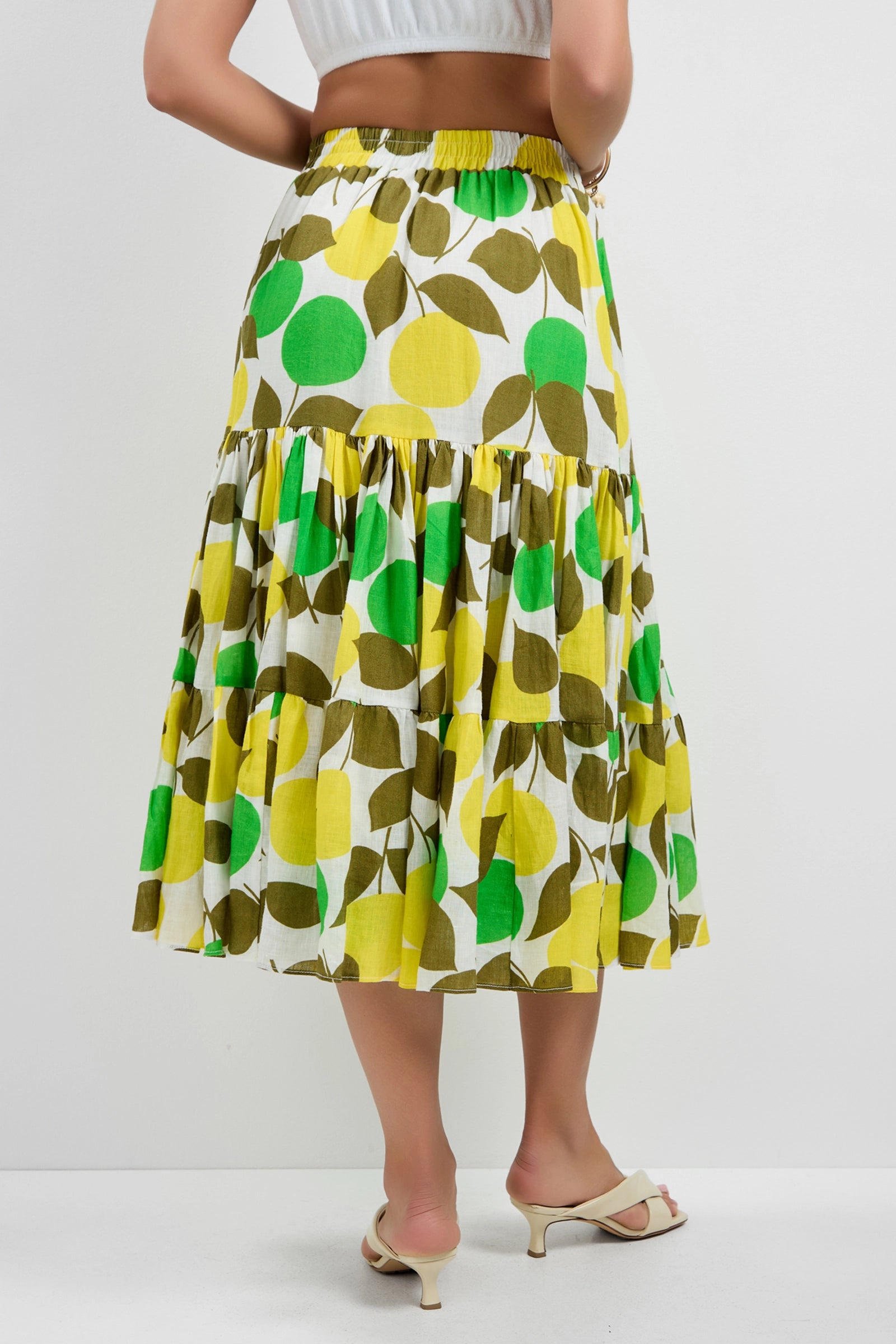 A woman stands against a plain white background wearing the Marsay Drop Waist Midi Skirt, featuring a white and green tiered design with a bold leaf pattern in green, yellow, and olive, paired with white heeled sandals.
